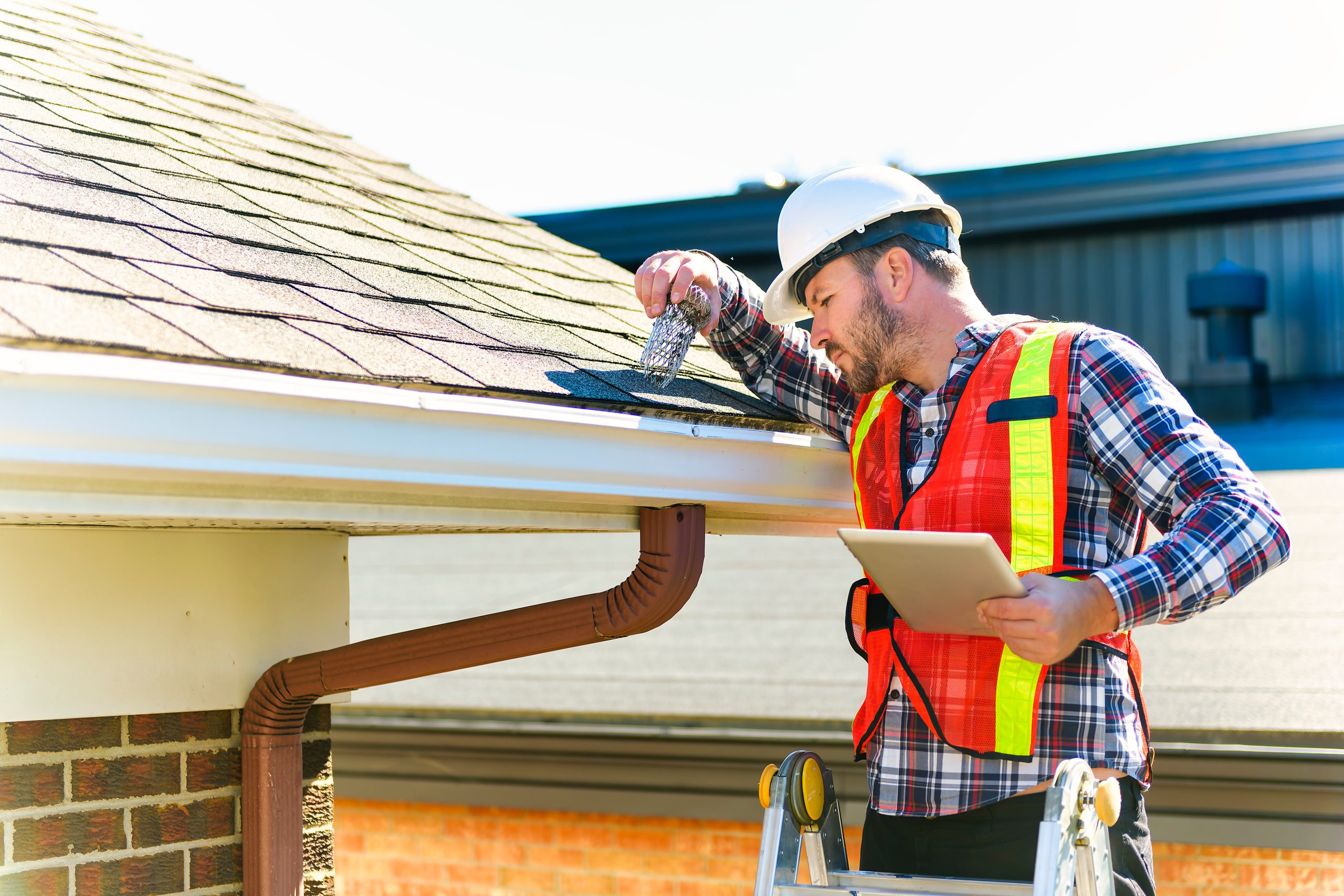man with hard hat standing on steps inspecting house roof Man in safety gear inspects a house roof and gutter while holding a tablet and standing on a ladder.