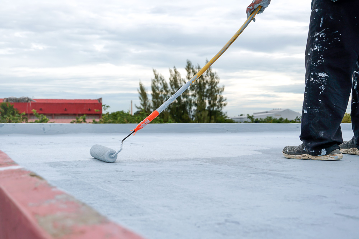 Person painting a flat rooftop with a roller brush under a cloudy sky.