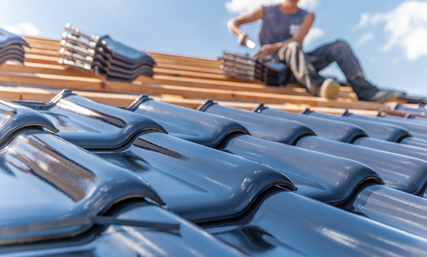 Close-up of shiny black roof tiles with a worker installing tiles on a roof in the background.
