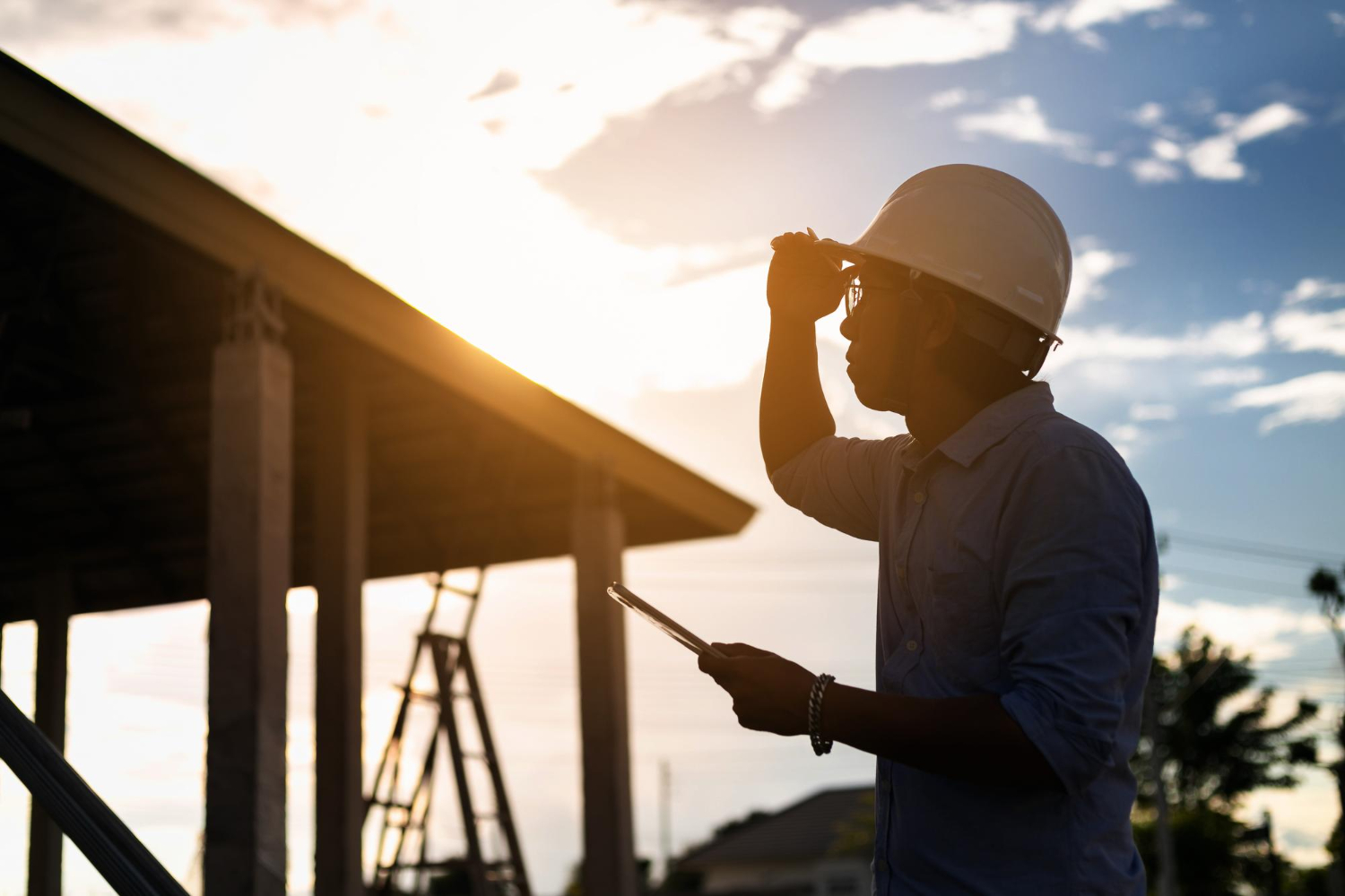 Silhouette of a construction worker with a hard hat holding a tablet at a building site during sunset.