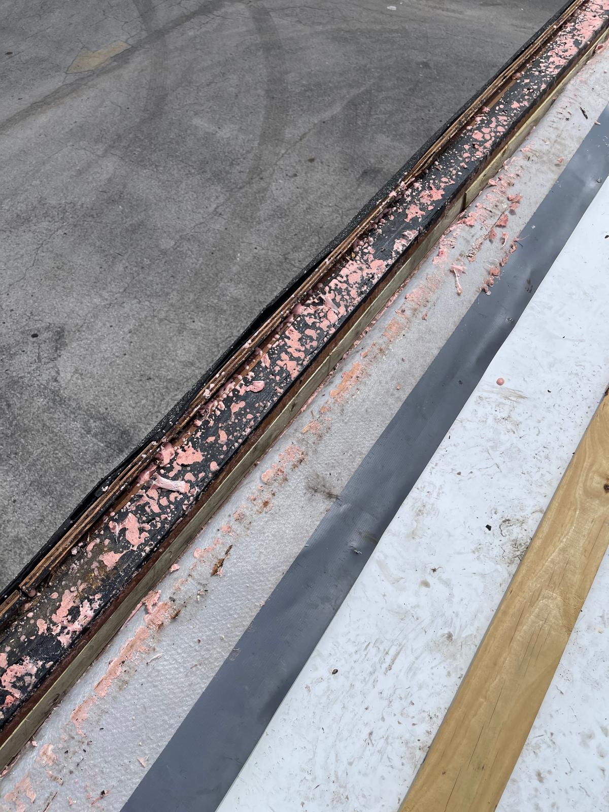 Close-up of a roof edge with exposed layers, pink sealant residue, and different construction materials.