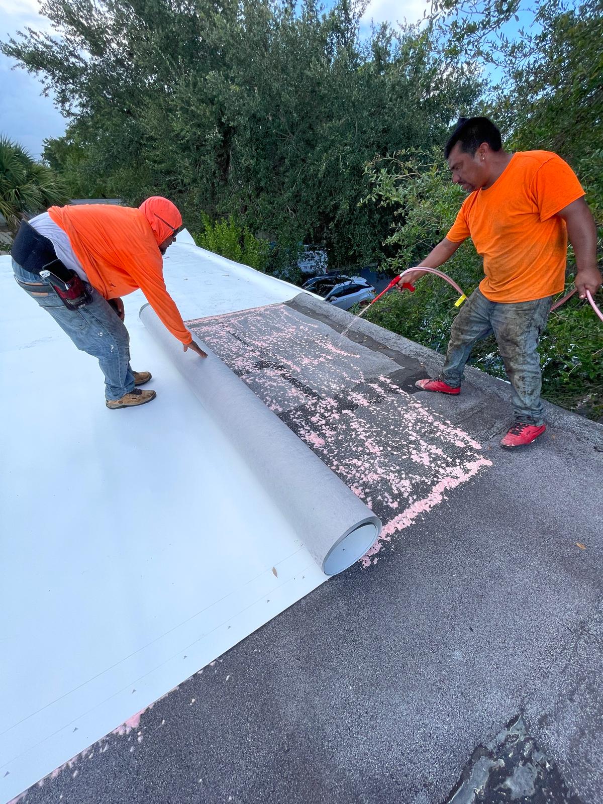 Two workers in orange shirts install roofing material, one rolling it out while the other sprays water.