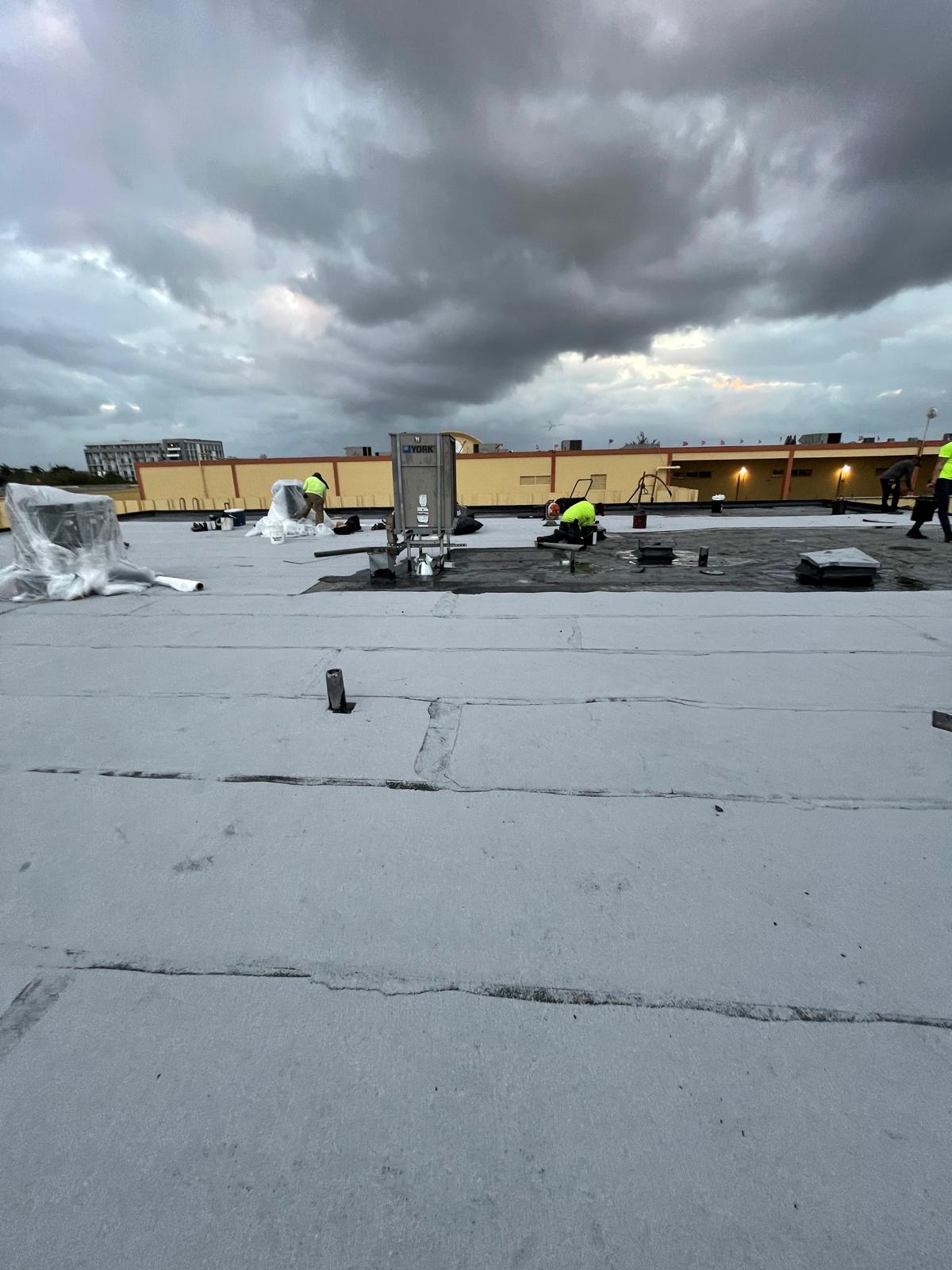 Two workers in safety vests work on a flat rooftop under a cloudy sky, with equipment and materials nearby.