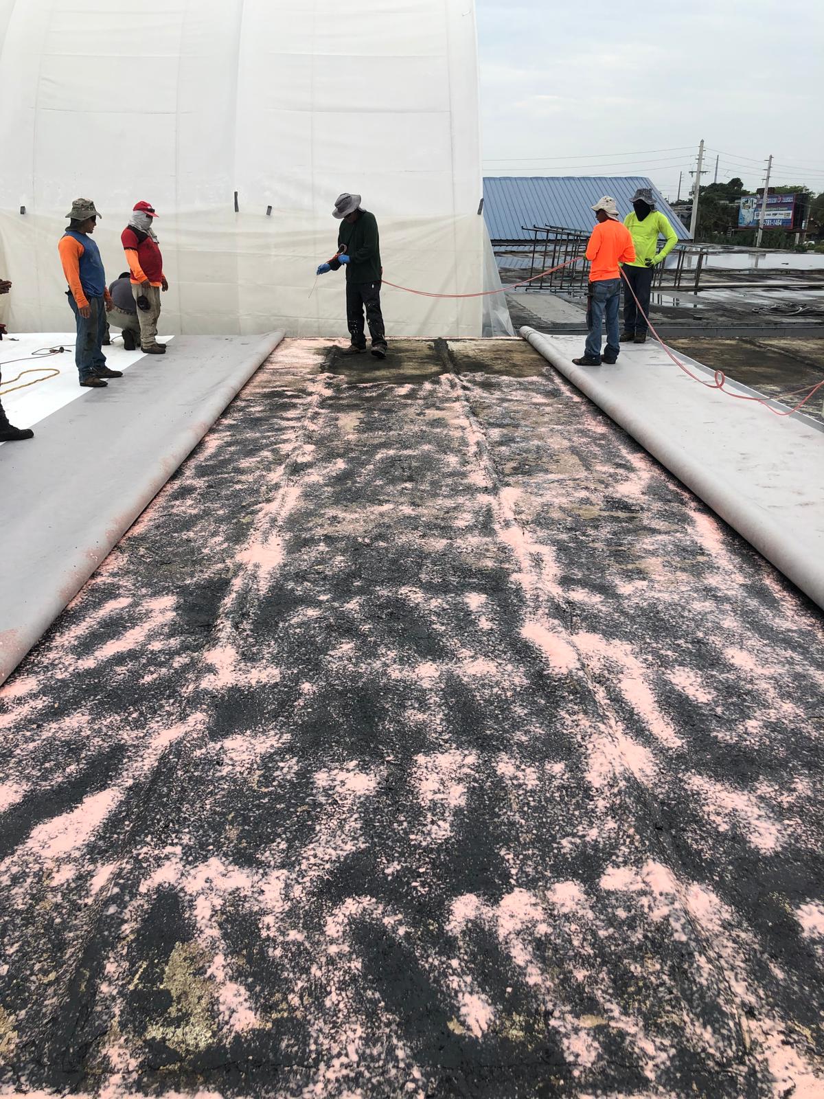 Several workers stand around a large rolled-out sheet on a construction site, with pink material visible on the surface.