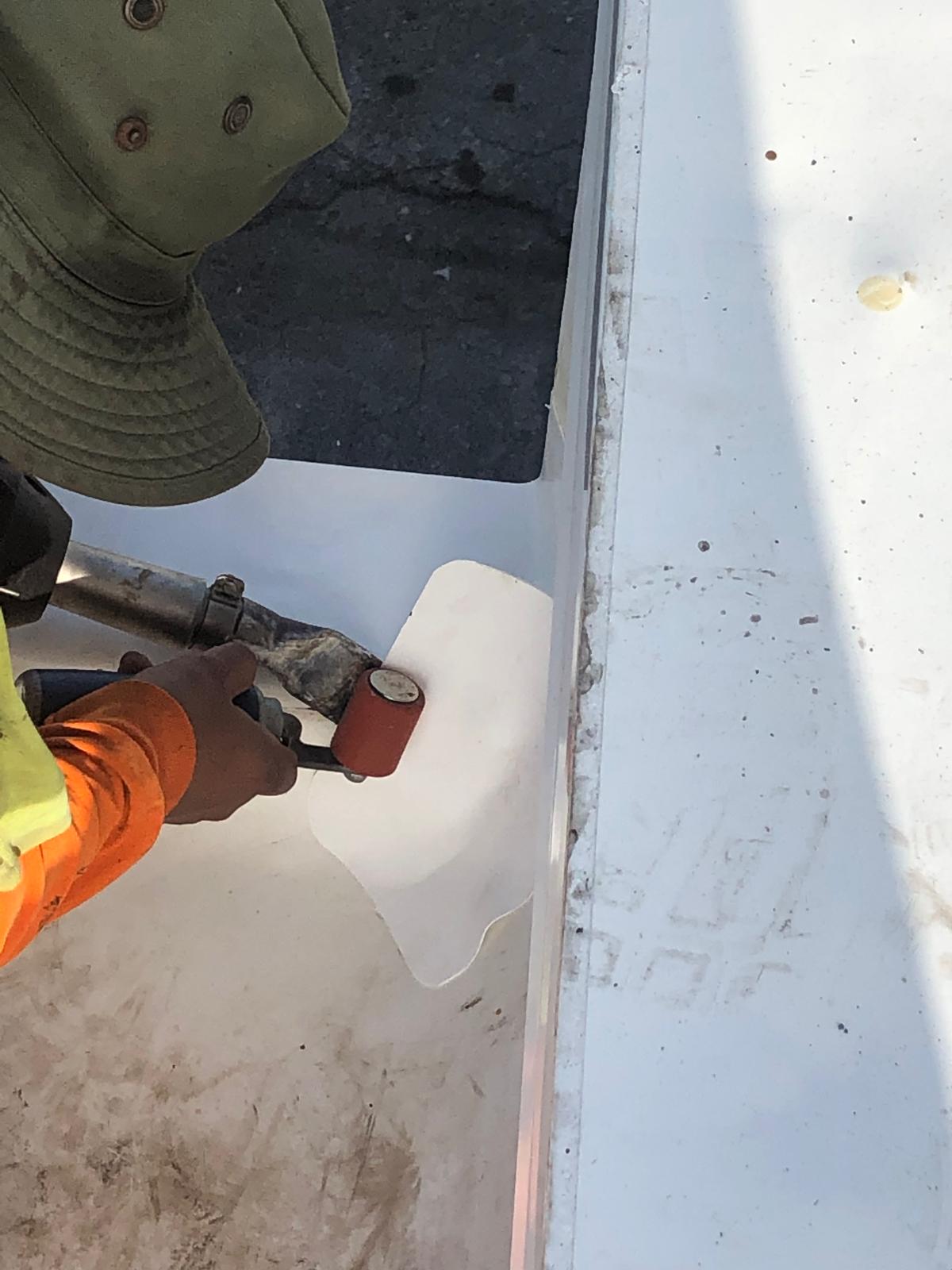 Person using a heat gun and roller to seal a white sheet onto a surface, wearing an orange sleeve and green hat.