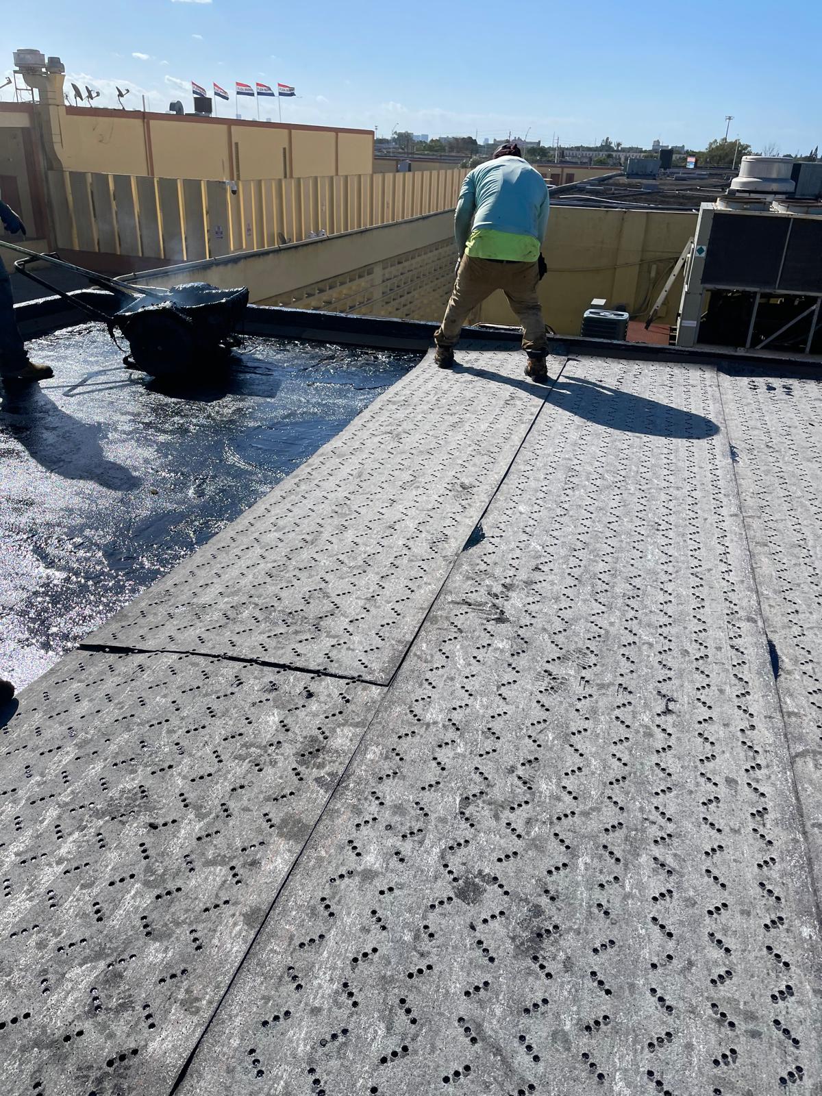 A worker installing roofing material on a flat building roof under clear blue sky, with tools nearby.
