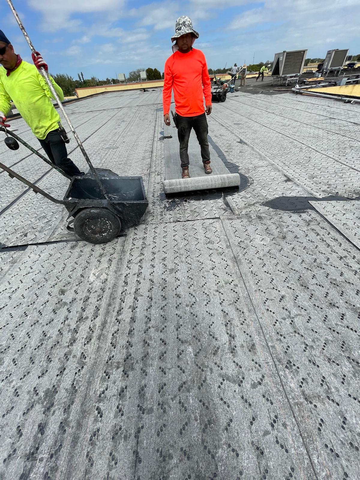 Two workers install roofing material on a flat rooftop under a partly cloudy sky.