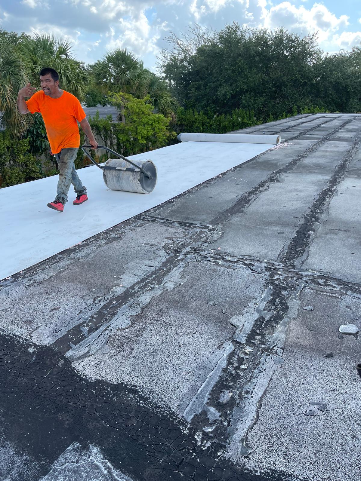 Man in orange shirt applies roofing material with roller on flat roof, surrounded by trees and greenery.