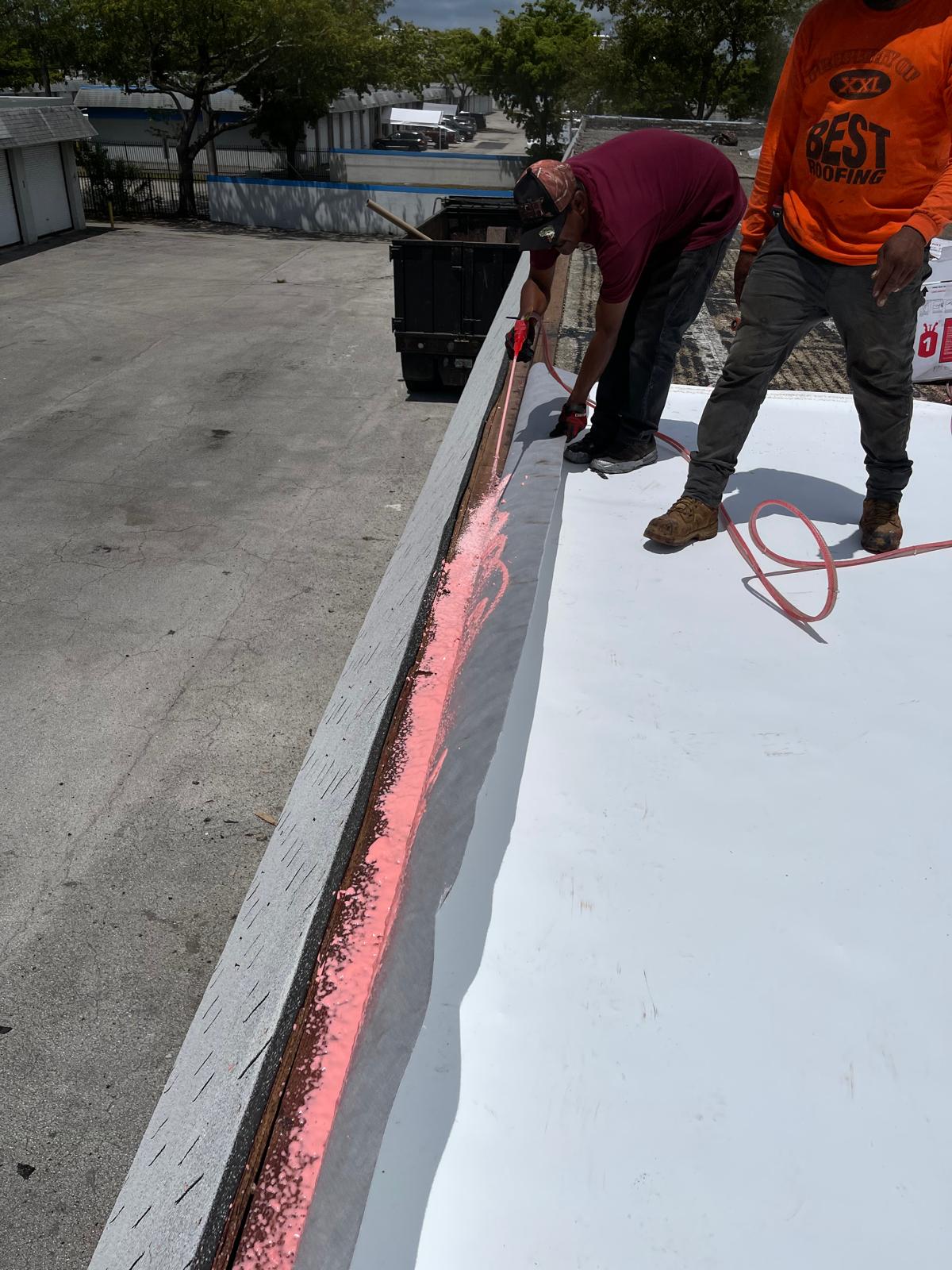 Two workers applying pink spray foam along the edge of a white rooftop under a partly cloudy sky.