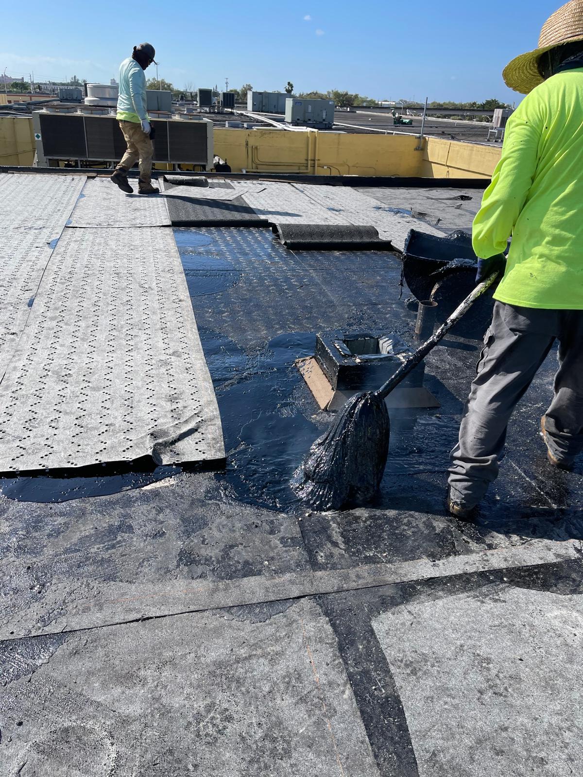 Two workers apply black waterproofing material to a flat rooftop under a clear sky.