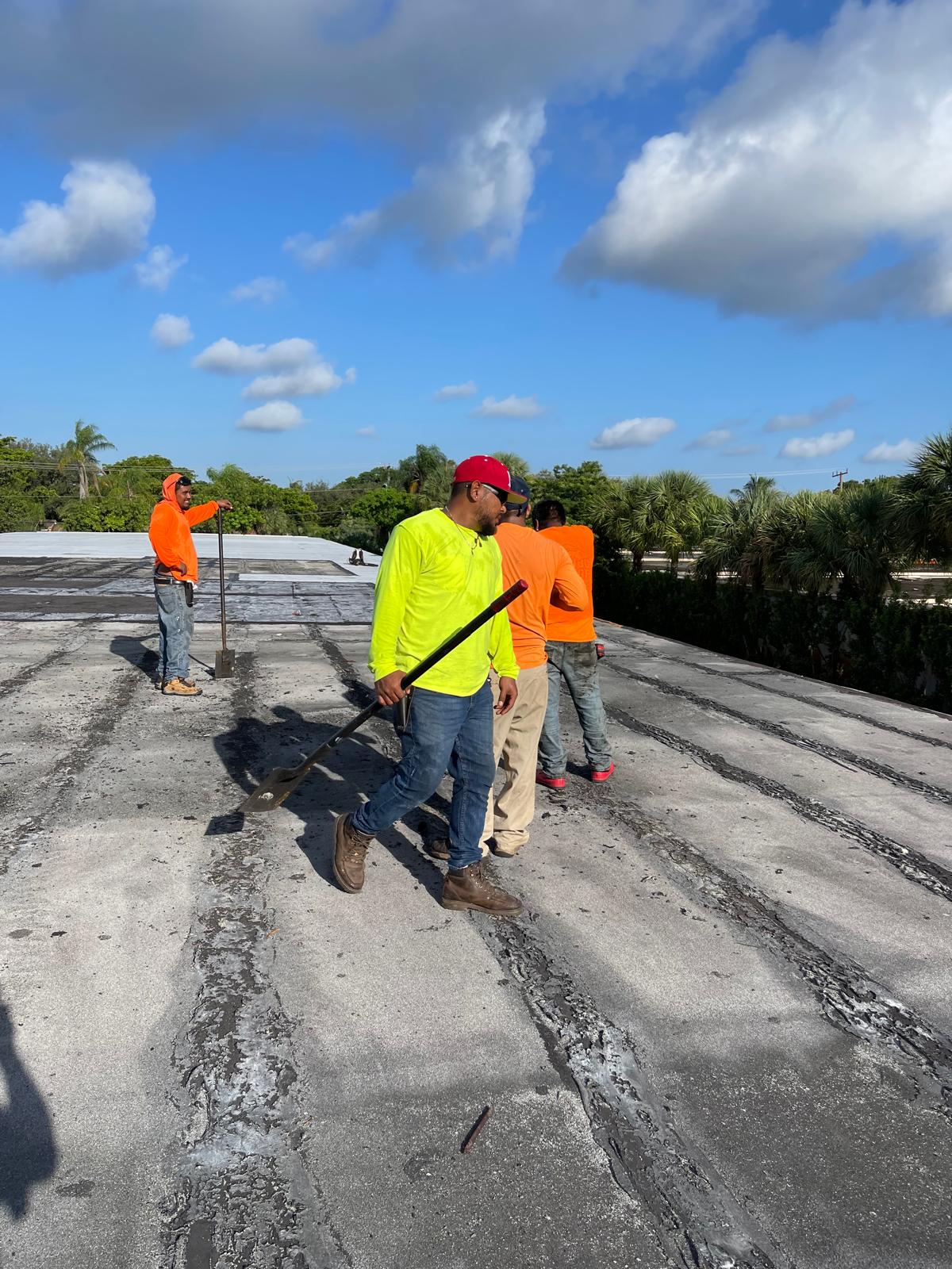 Four workers in bright shirts cleaning or repairing a flat rooftop under a blue sky with scattered clouds.