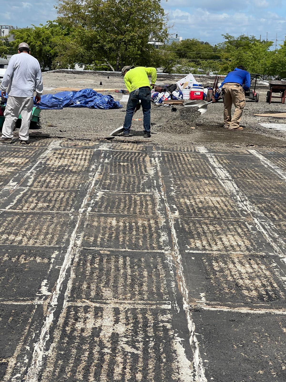 Three workers repair or clean a weathered, stripped roof under a sunny sky with tools and supplies nearby.