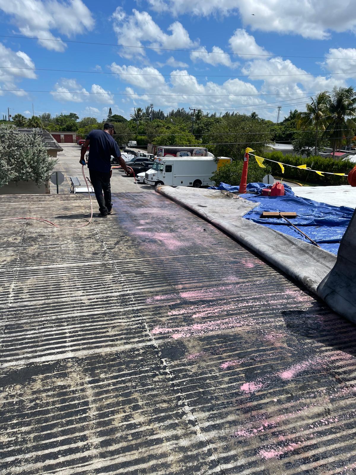 Worker spraying material on a rooftop under sunny skies, with vehicles and trees in the background.