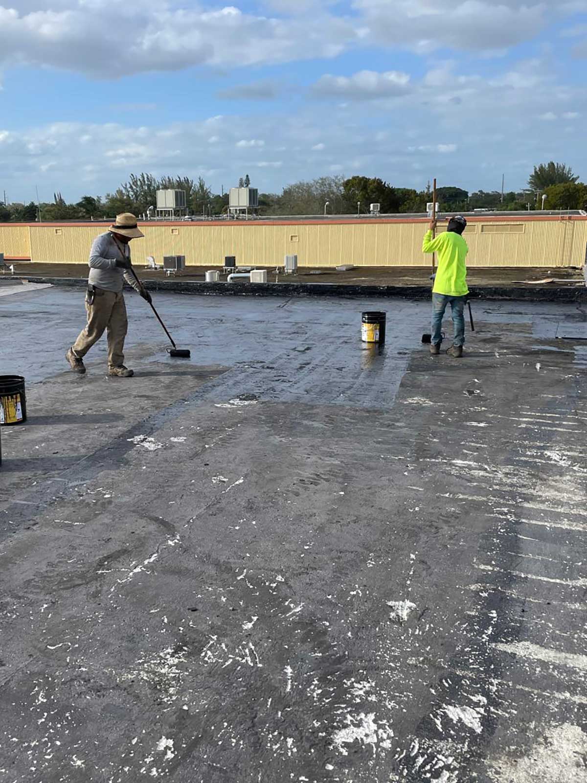 Two workers apply black sealant to a flat rooftop under a partly cloudy sky, with tools and buckets nearby.