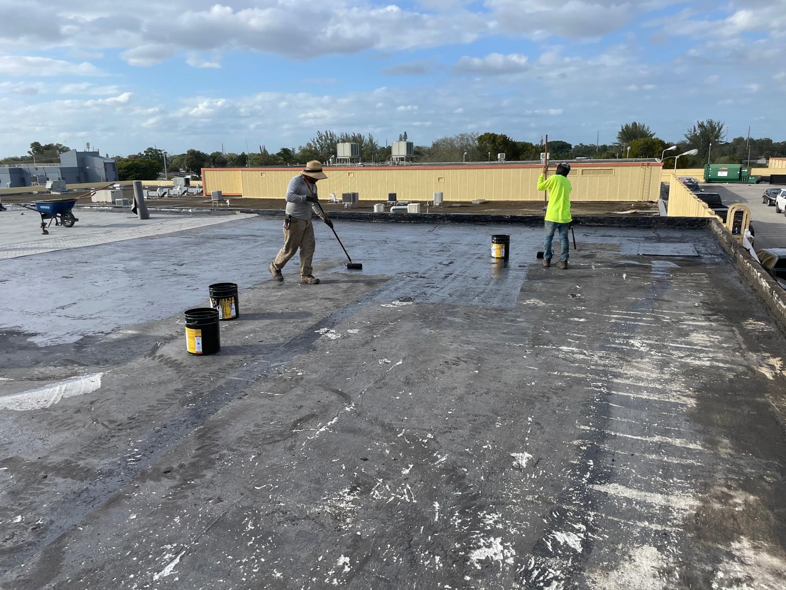 Two workers apply coating to a flat rooftop with buckets and rollers under a partly cloudy sky.