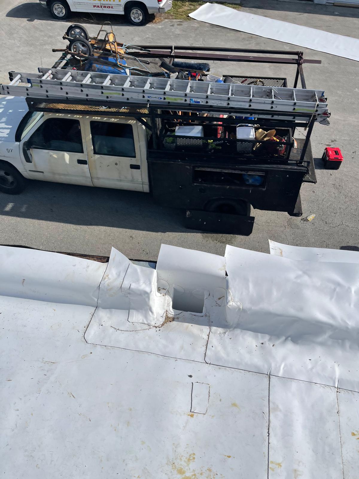 A work truck with ladders and tools is parked next to a building with a white, patched roof.