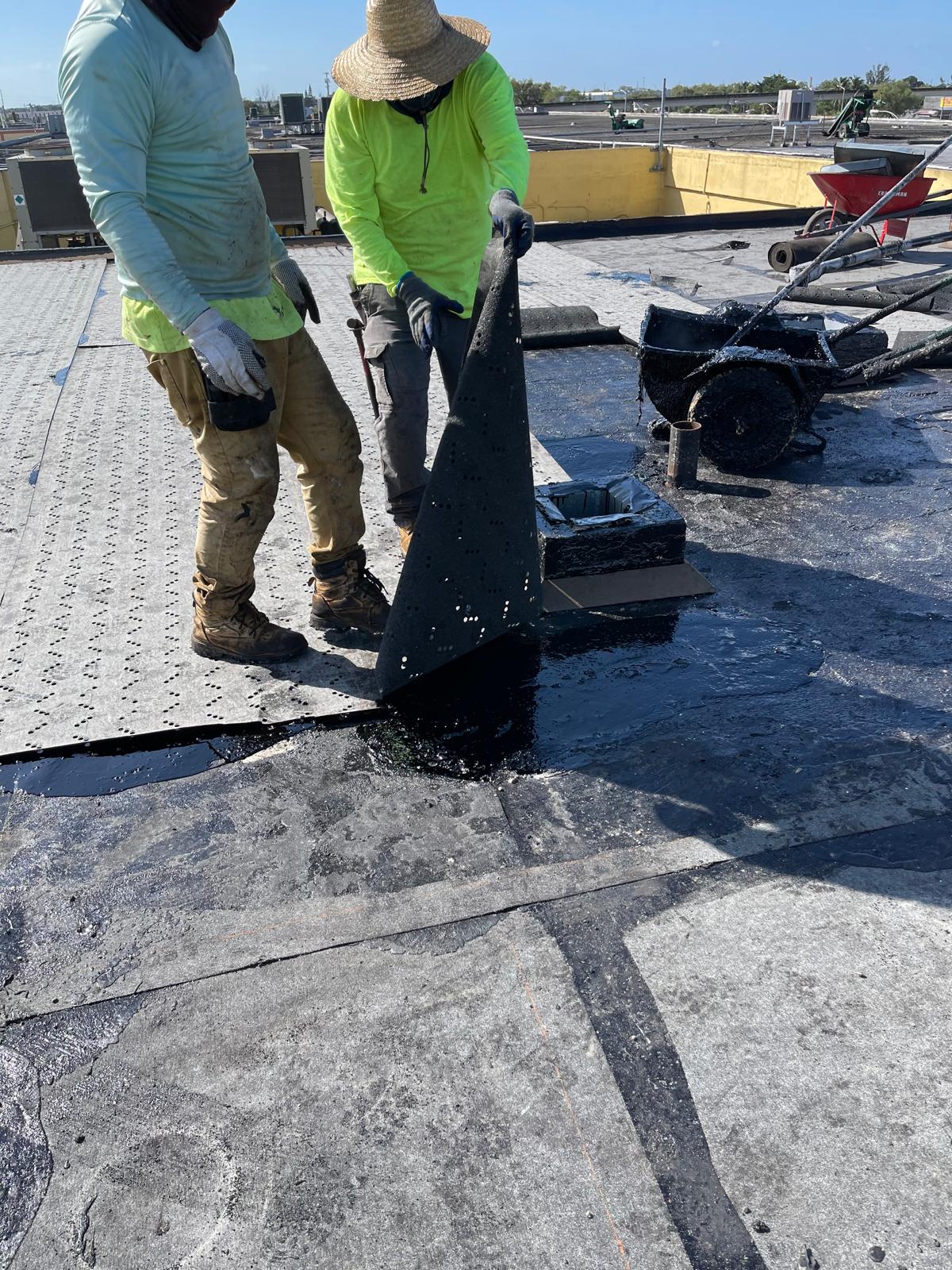 Two workers in protective clothing install roofing material on a flat roof under sunny skies.