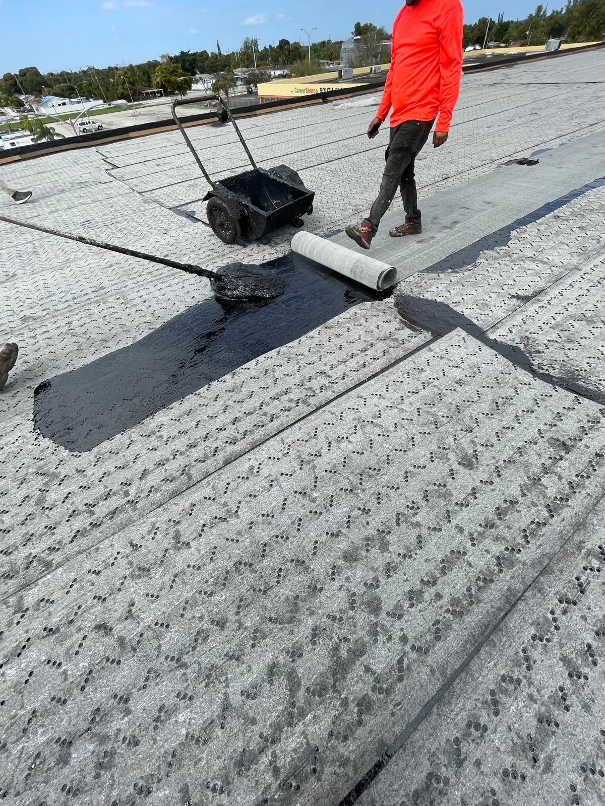 Worker applies black sealant and rolls out roofing material on a flat rooftop under a cloudy sky.
