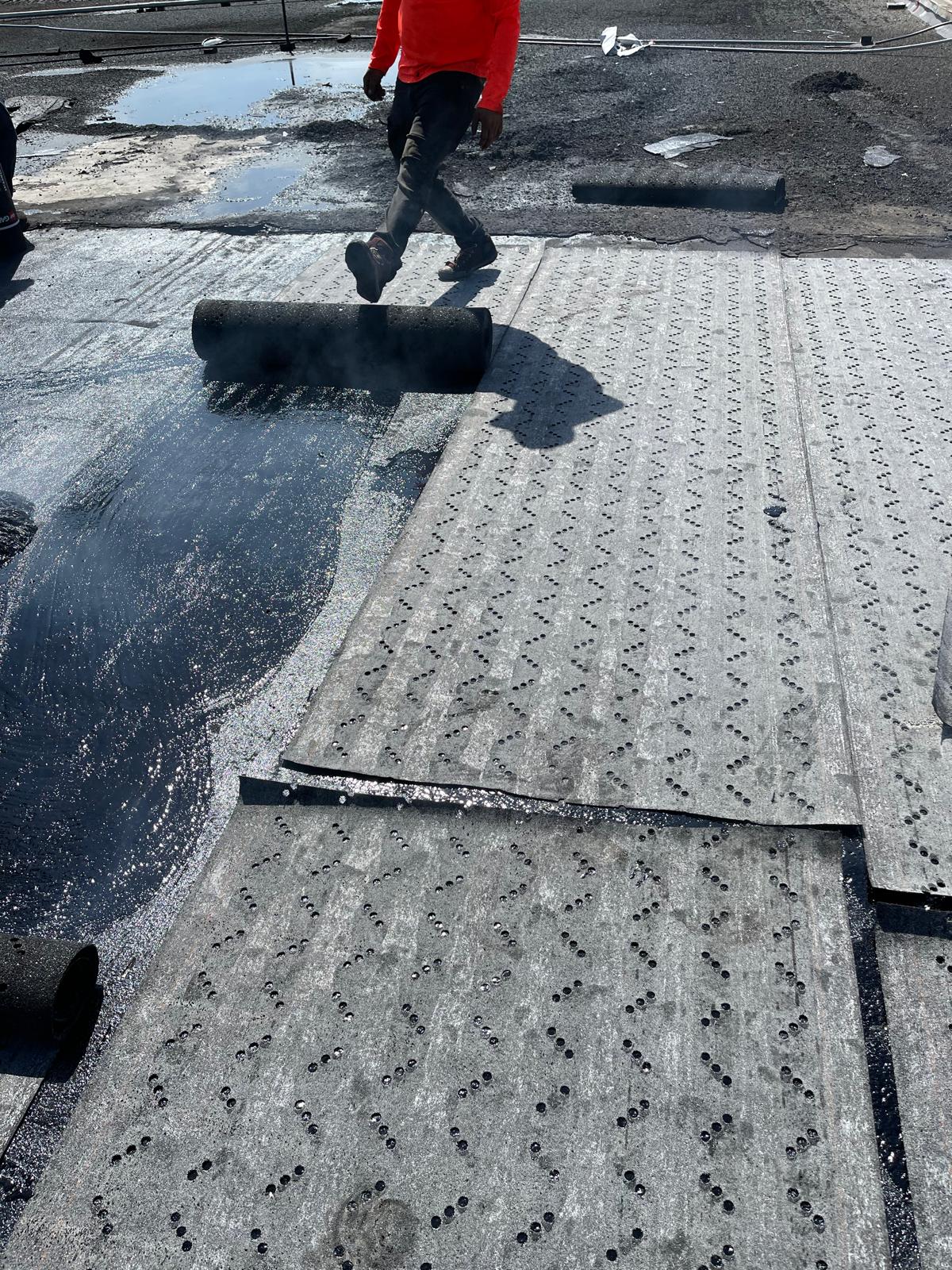 Worker in a red shirt installing roofing material on a flat roof with black rolls and tar visible.