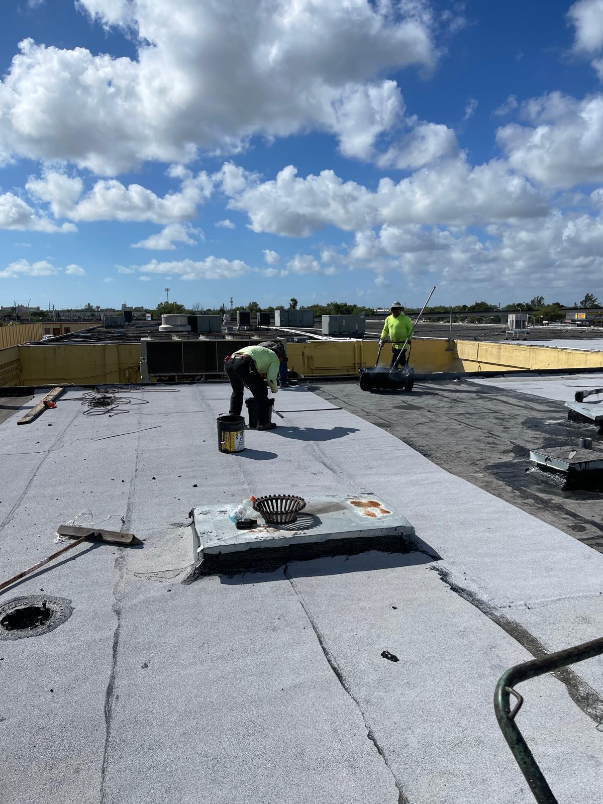 Two workers in safety gear install roofing material on a flat roof under a partly cloudy sky.