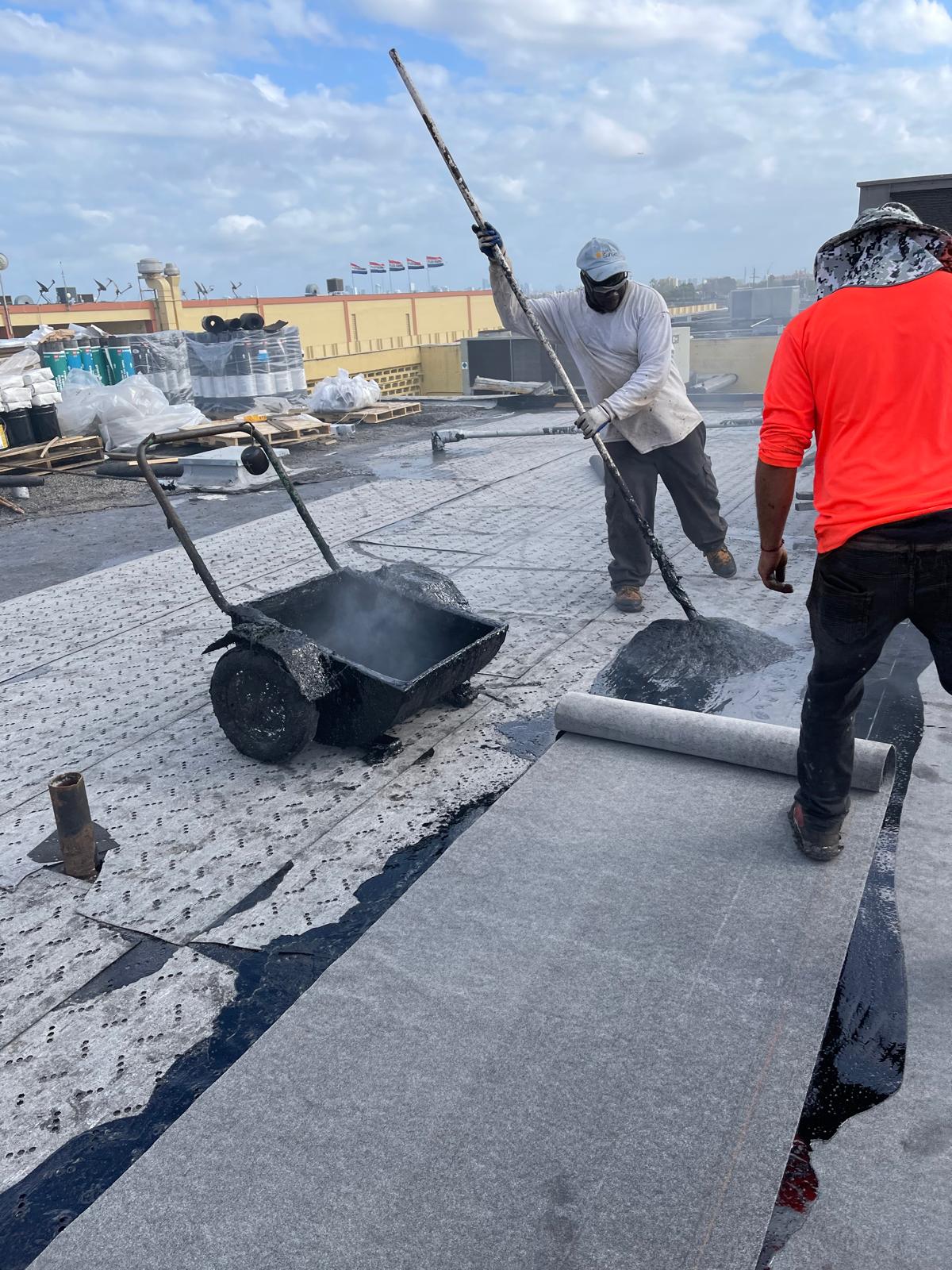 Two workers apply roofing materials on a flat roof under a partly cloudy sky, with equipment and supplies nearby.