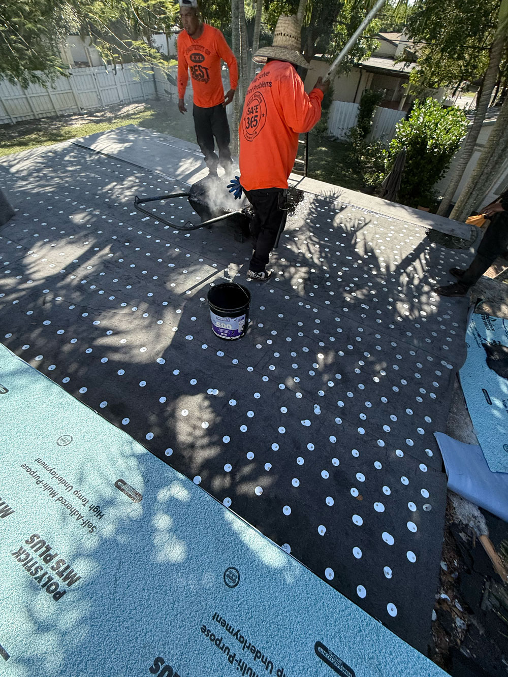 Roofers in orange shirts install waterproof underlayment with adhesive dots and tools on a sunny day.