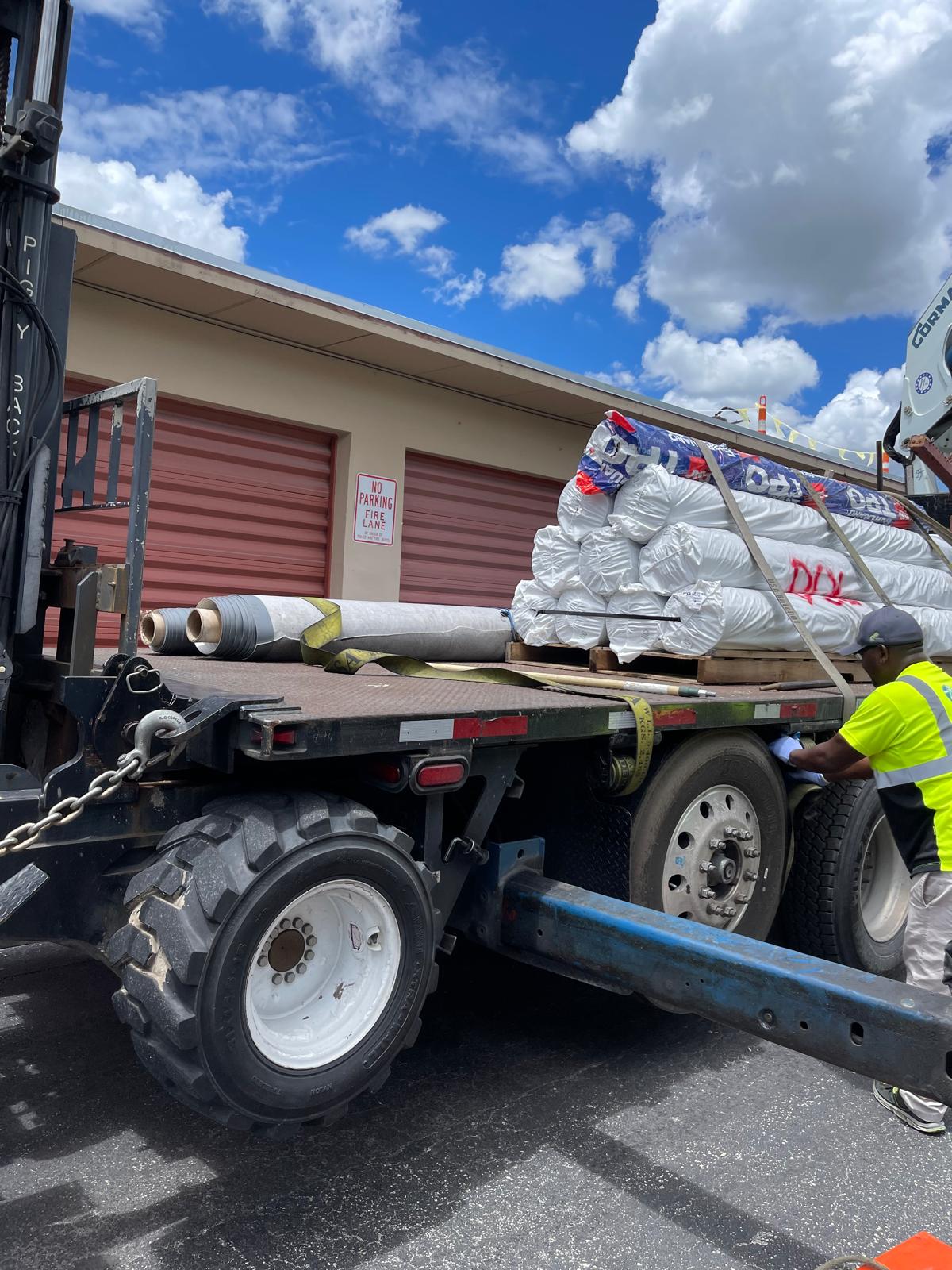 A worker secures rolled materials on a flatbed truck near storage units under a partly cloudy sky.