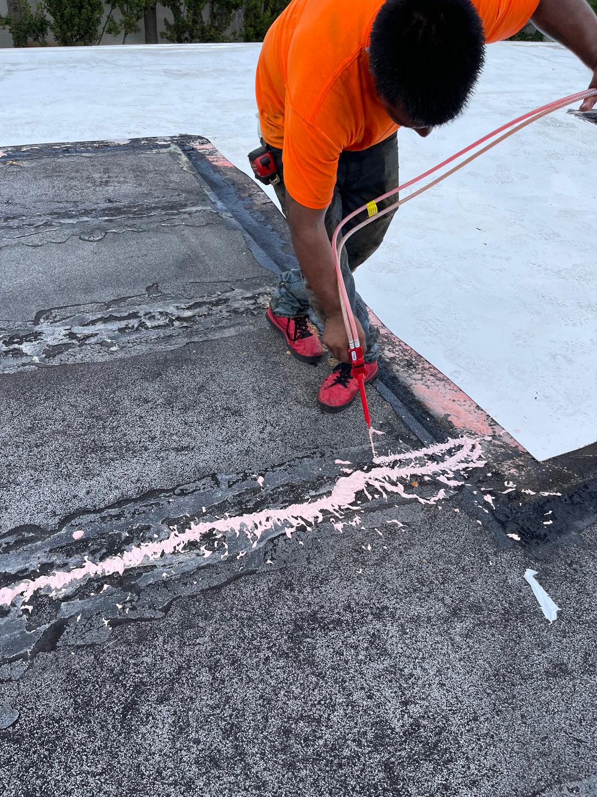 Person in orange shirt applies pink sealant to a crack on a flat, weathered roof using a dispensing tool.