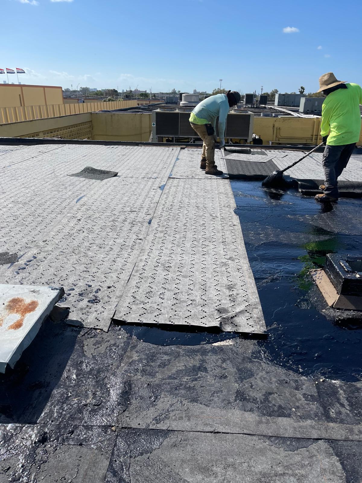 Two workers in hats install roofing material on a flat roof under a clear blue sky.