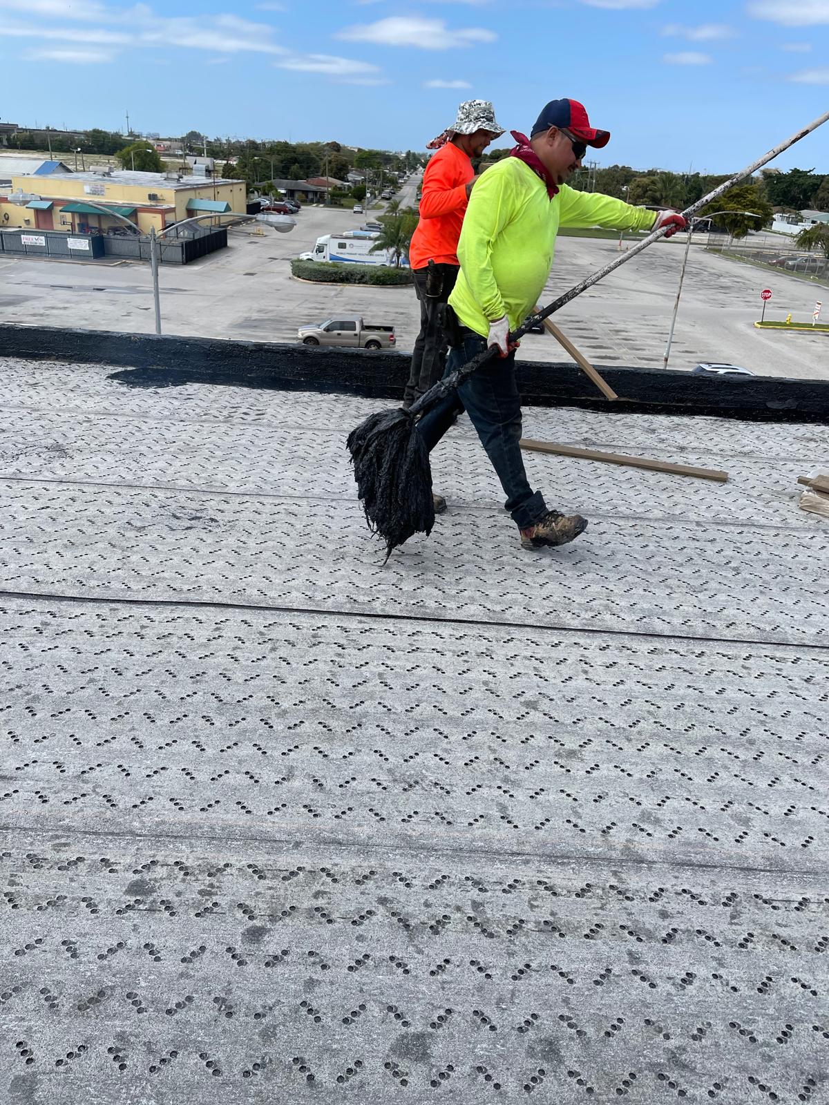 Two workers on a rooftop spread tar or asphalt with tools on a sunny day, with buildings visible in the background.