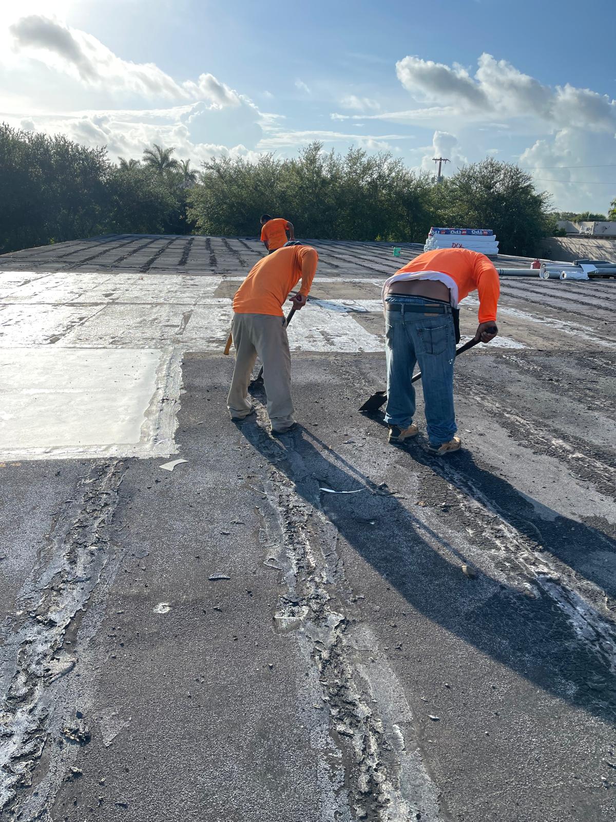 Two workers in orange shirts repair a flat rooftop under a bright blue sky with scattered clouds.