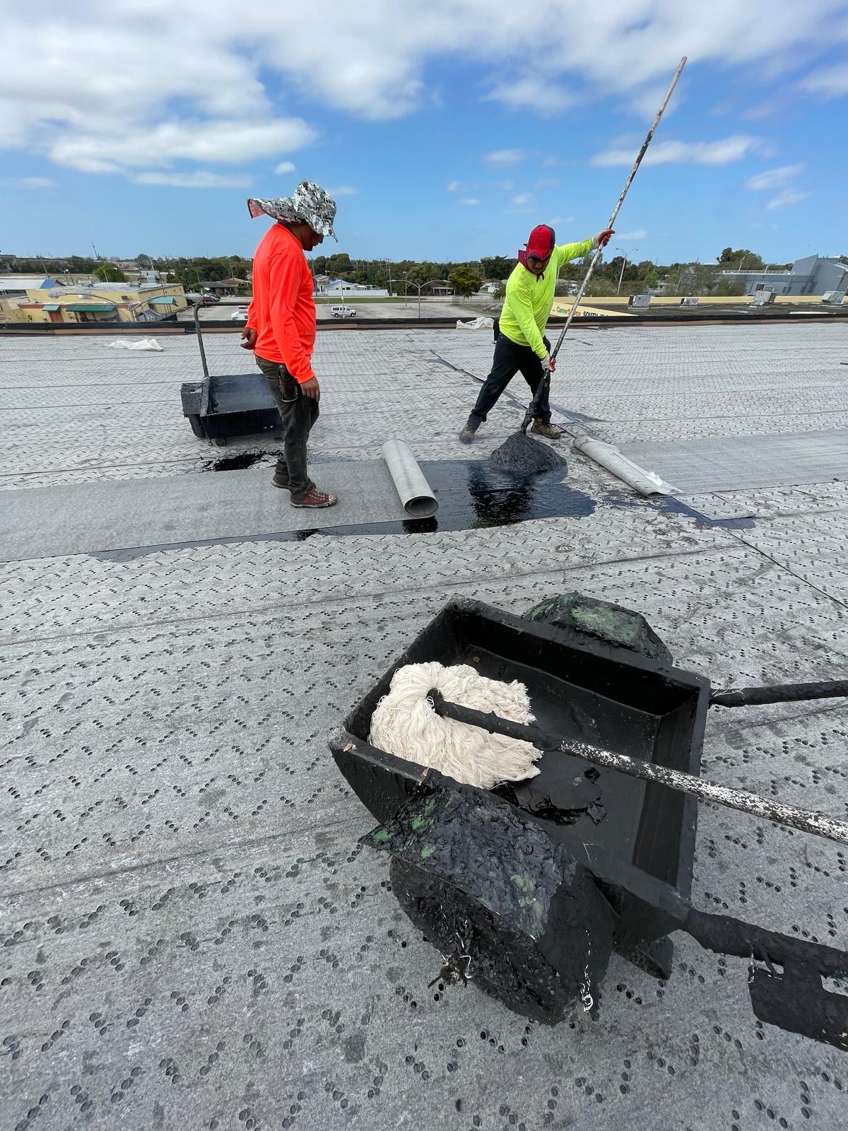Two workers in bright shirts apply black sealant to a flat rooftop under a partly cloudy sky.