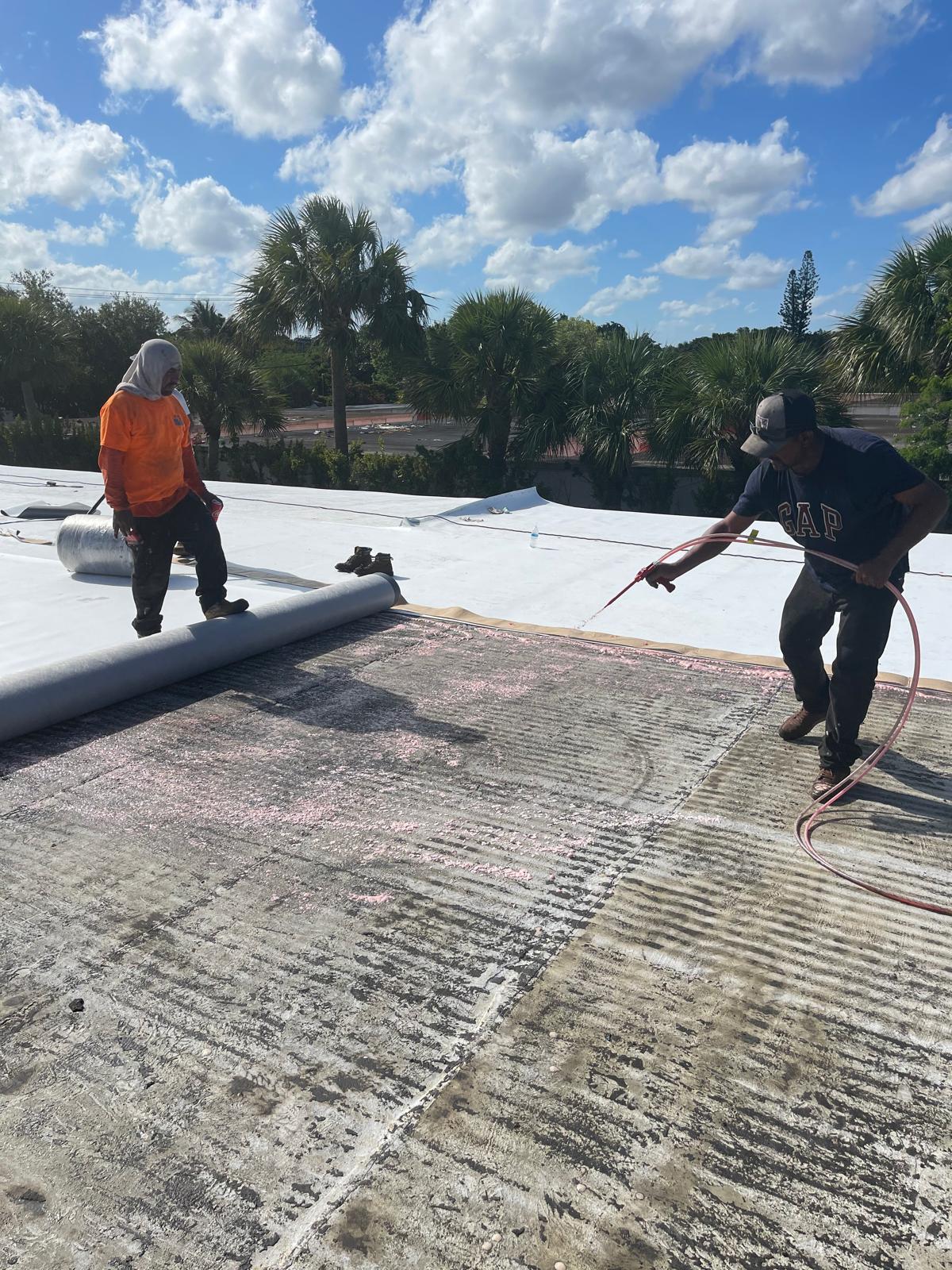 Two workers repair a rooftop, one spraying and one standing with roofing material under a sunny sky.