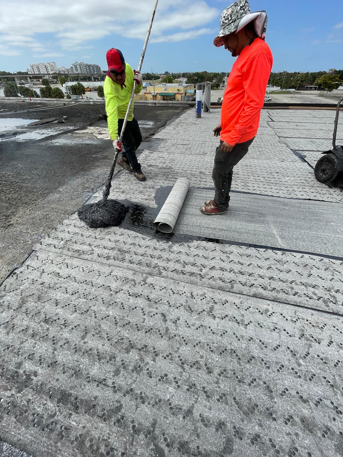 Two workers in bright shirts apply black material and unroll roofing felt on a flat rooftop under blue sky.