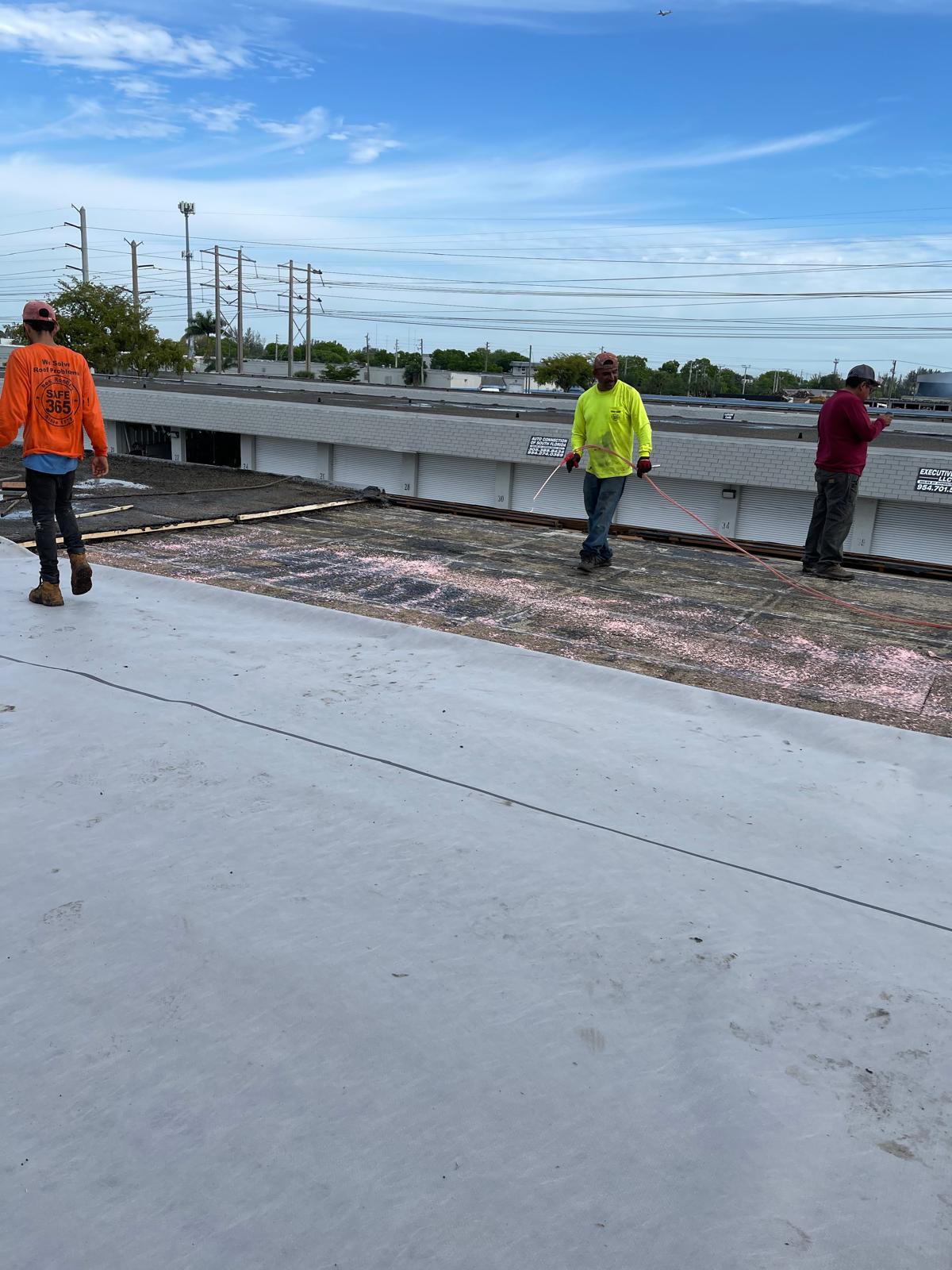 Three construction workers stand and walk on a flat rooftop under a blue sky with power lines in the background.