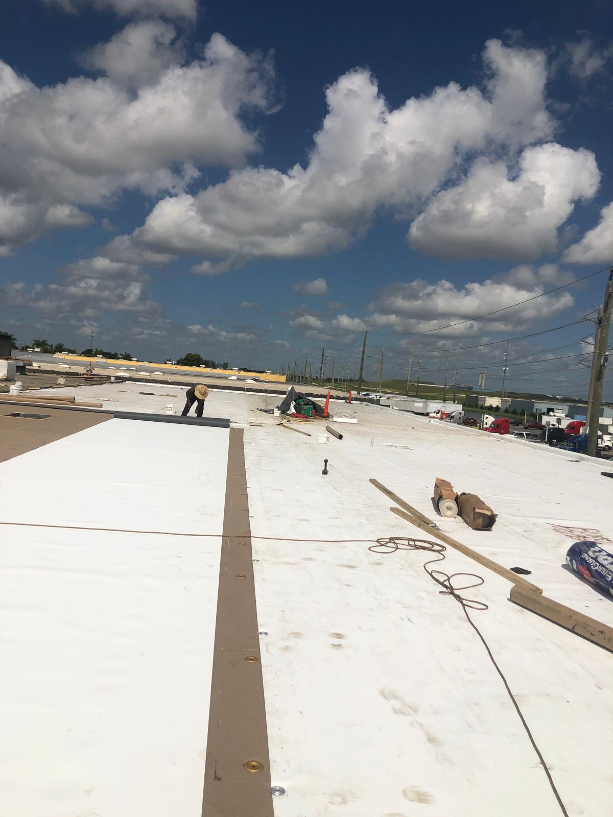 Workers installing roofing materials on a large flat roof under a partly cloudy sky.