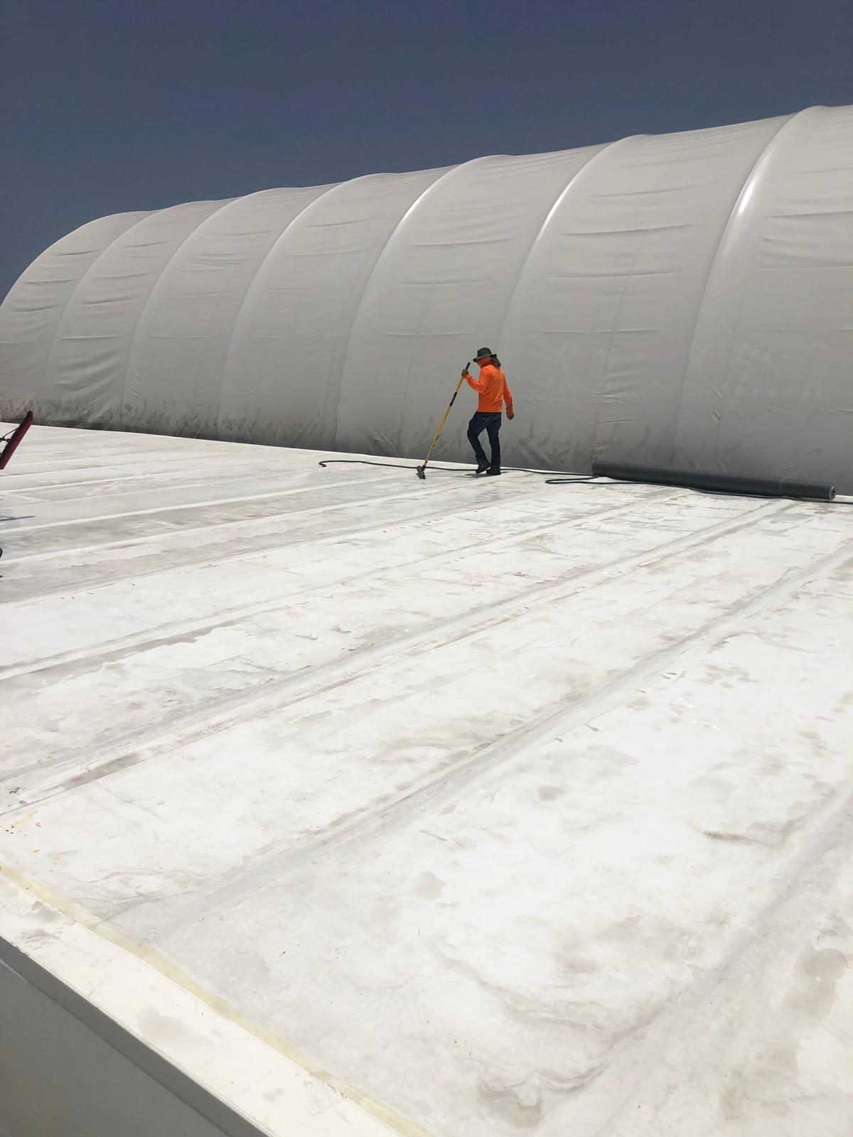 Worker in orange vest cleaning a large white rooftop near an inflatable white structure under clear sky.