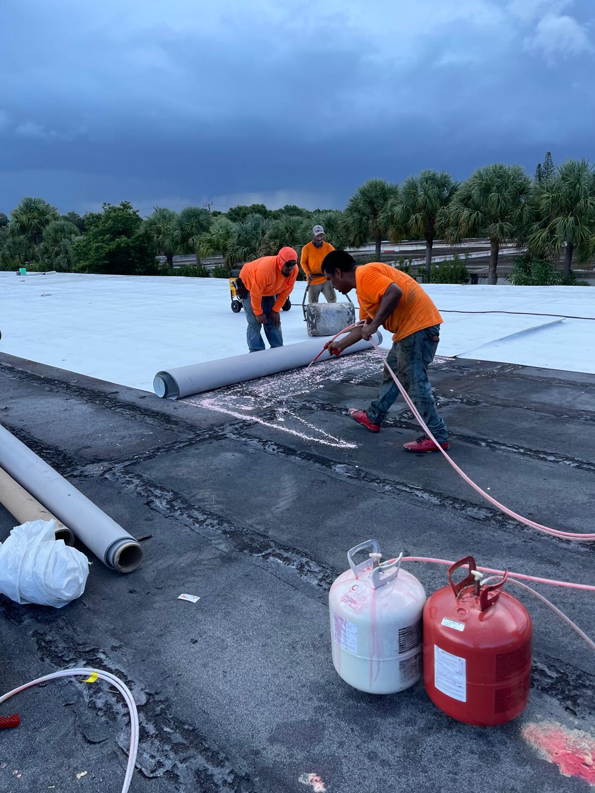 Three workers in orange shirts roll out roofing material and use a torch on a flat roof under a cloudy sky.