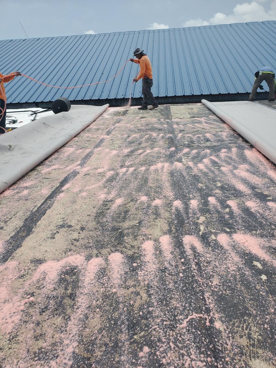 Two workers in orange shirts repair a large blue metal roof, spreading material and using safety ropes.