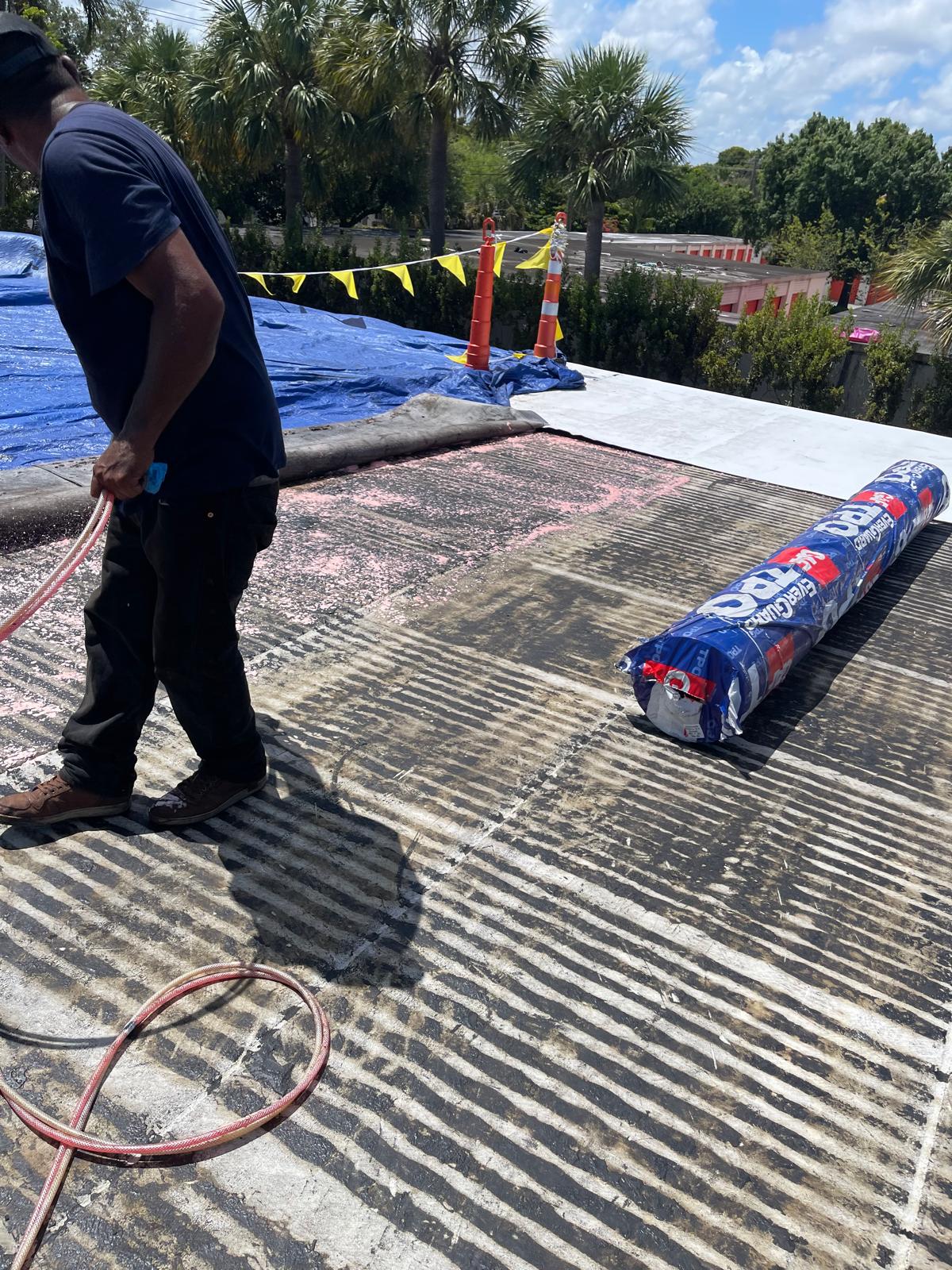 Worker prepares roofing surface with rolled material on a sunny day; palm trees and cones in background.