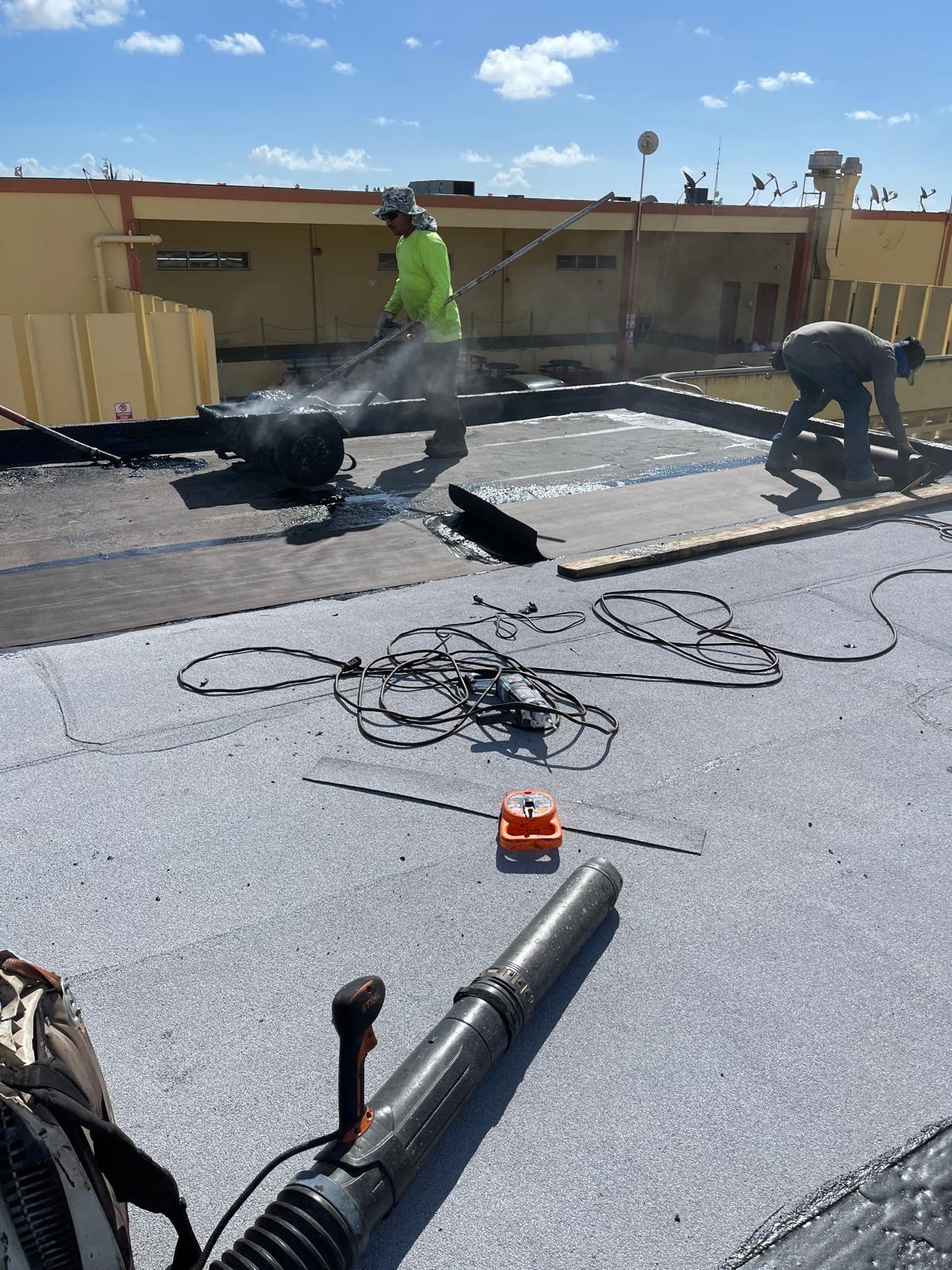 Two workers apply roofing material on a flat roof; tools and cables are scattered in the foreground.