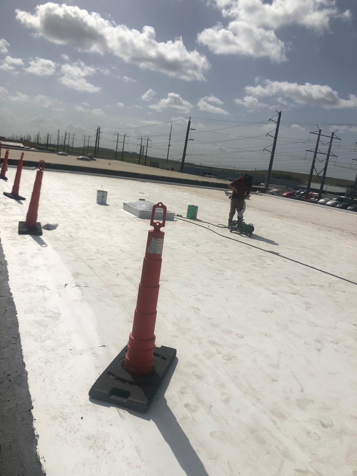 Person working on a flat white rooftop, surrounded by orange safety cones, under a partly cloudy sky.