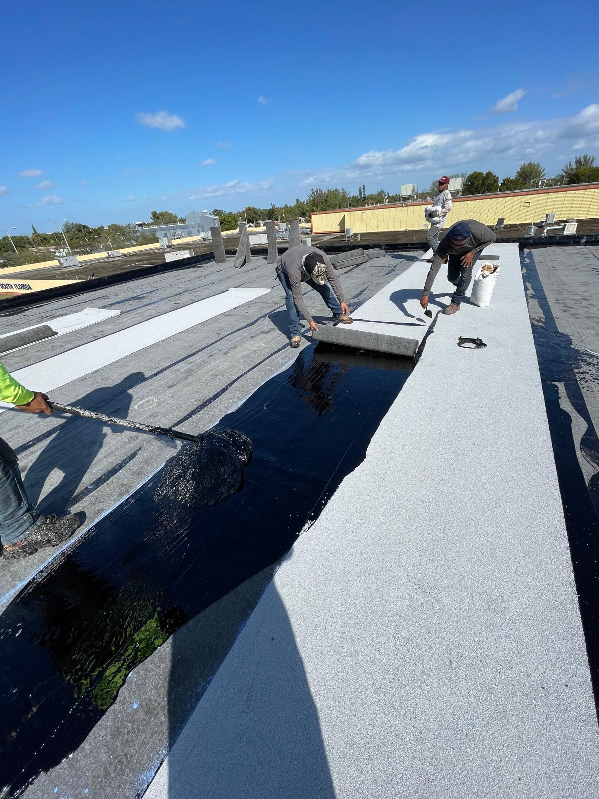 Workers installing roofing materials on a flat rooftop under a clear blue sky.