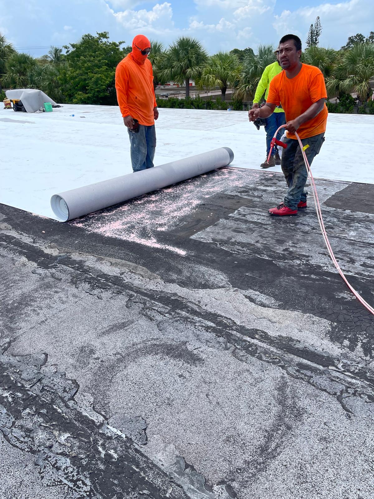 Two workers in orange shirts unroll roofing material on a flat roof under a partly cloudy sky.