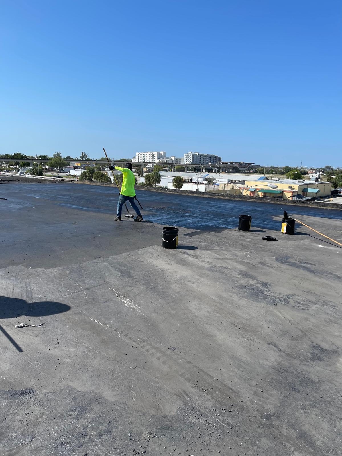 A worker in a neon shirt applies material to a flat rooftop under a clear blue sky.
