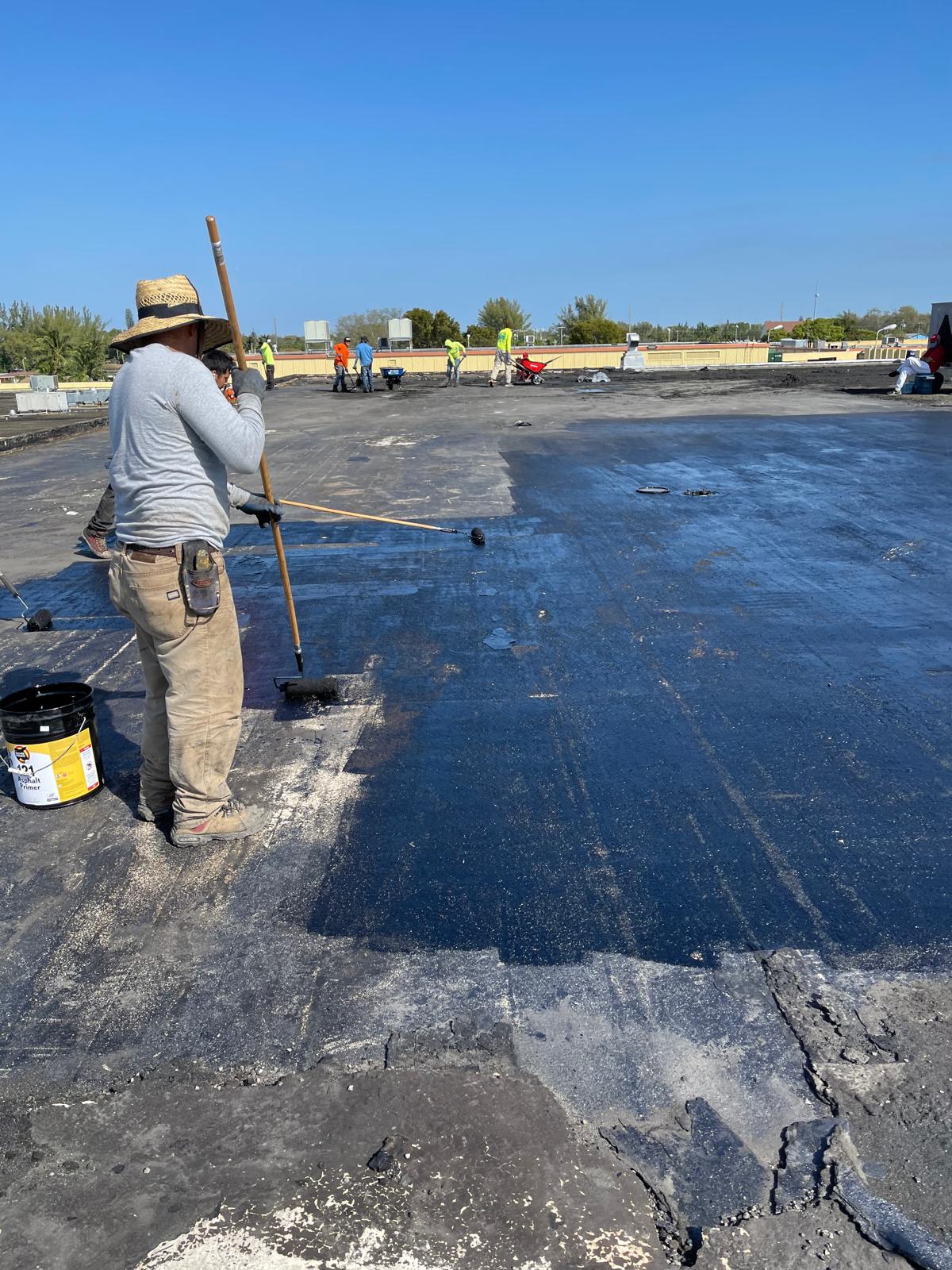 A construction worker applies black sealant to a flat rooftop; more workers are visible in the background.