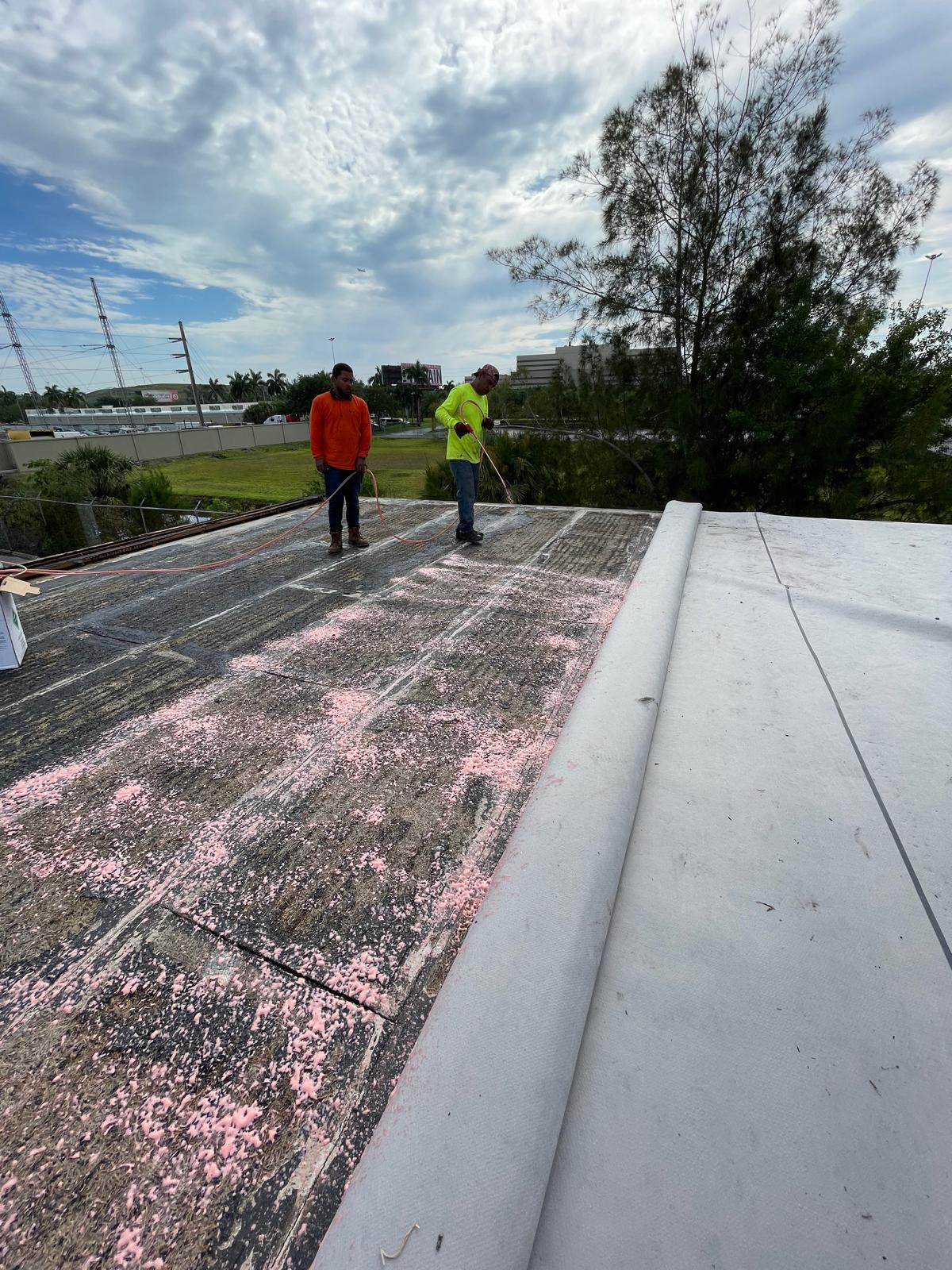 Two workers prepare and clean a flat rooftop, rolling out white roofing material under a cloudy sky.