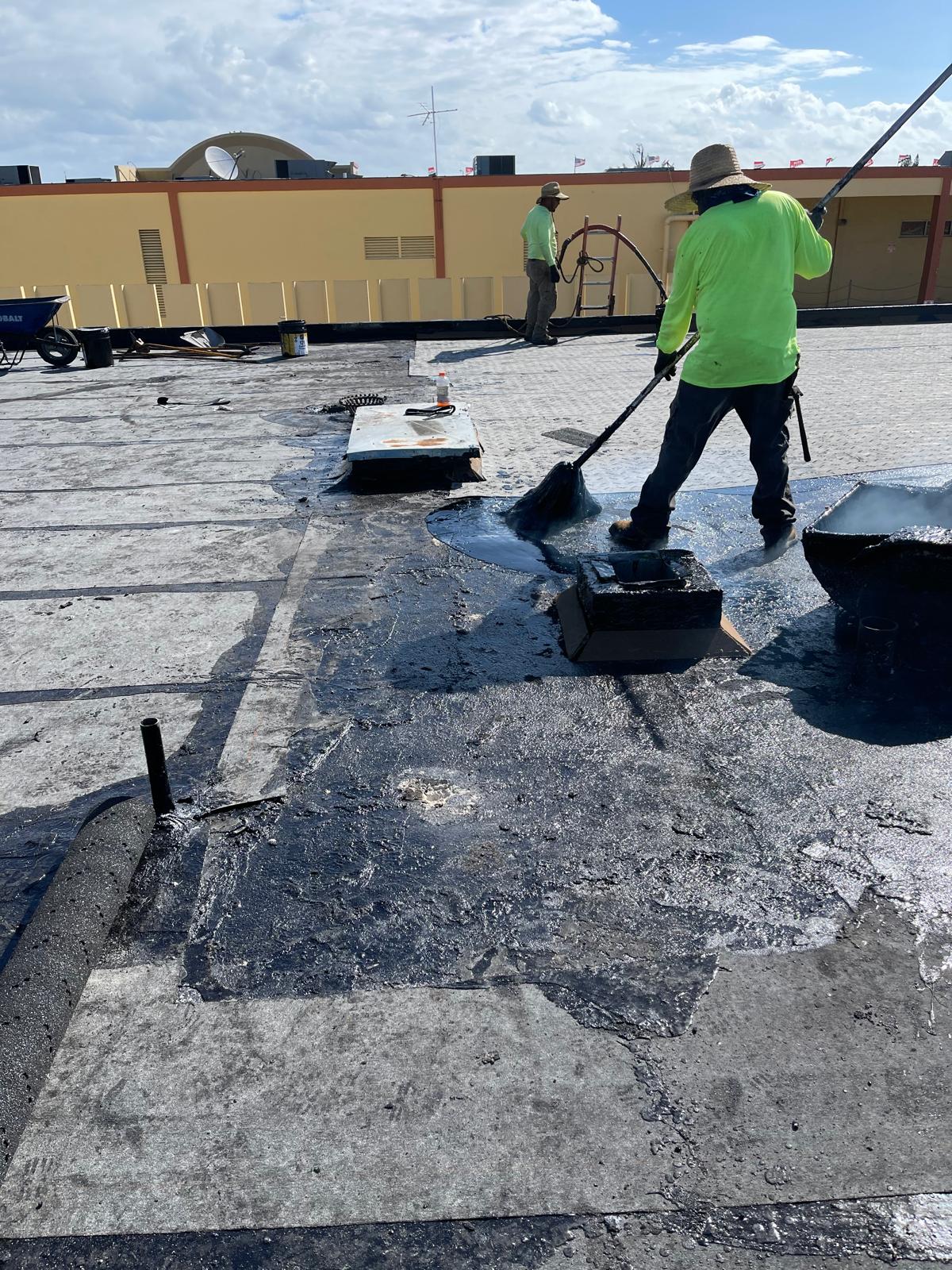 Two workers in hats and neon shirts apply roofing material with brooms on a flat rooftop under a partly cloudy sky.