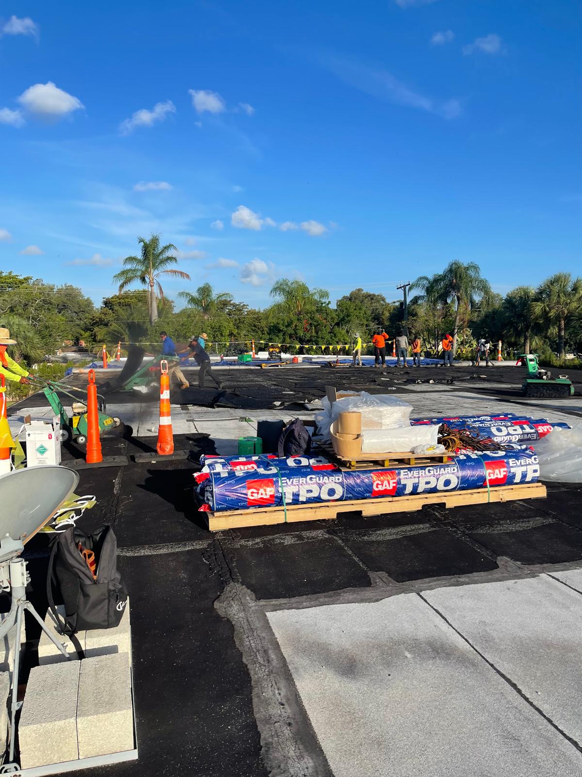 Roofers working on a flat roof with construction materials and palm trees in the background under a blue sky.