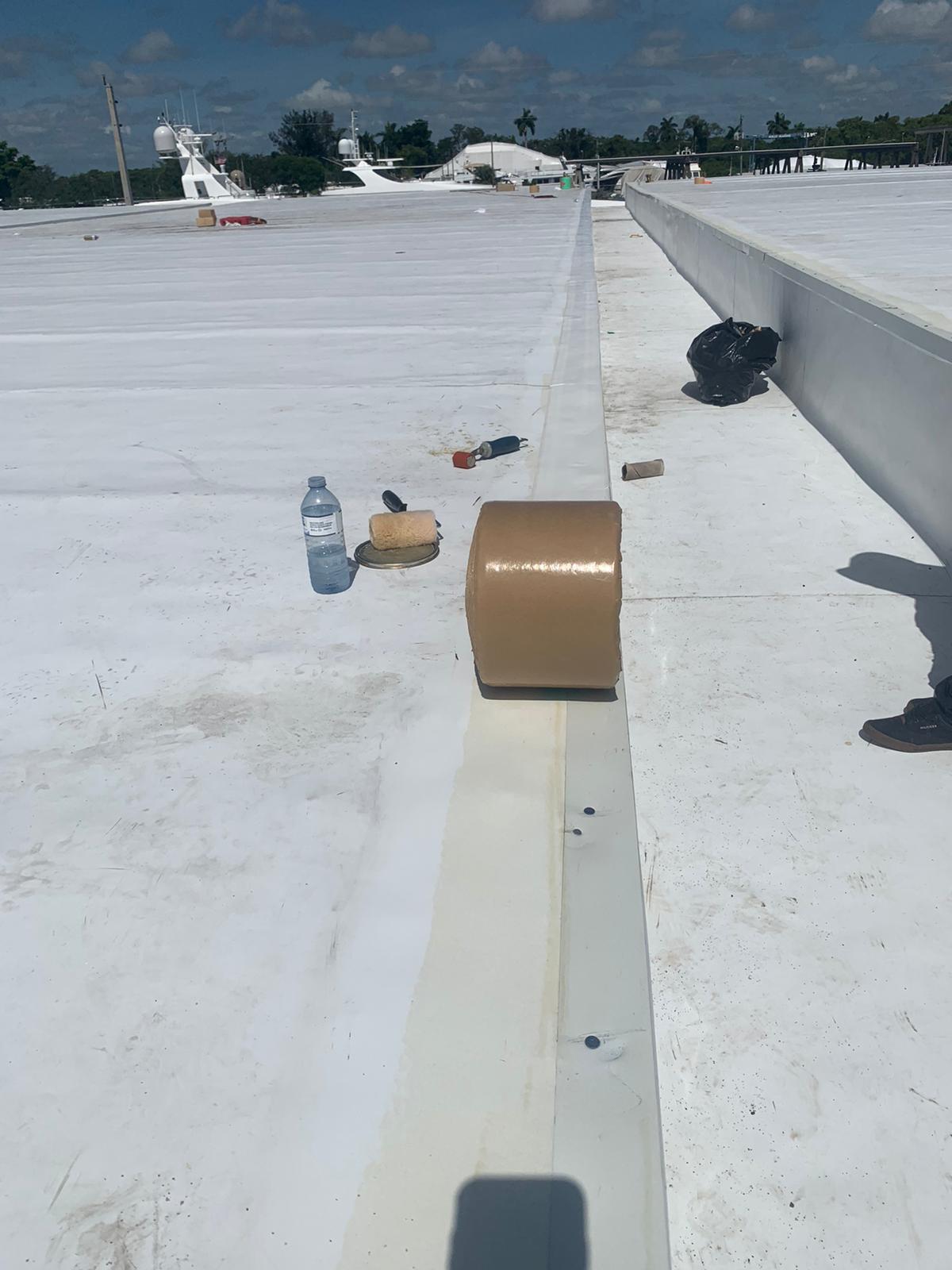A roll of brown tape sits on a white rooftop with tools and a water bottle nearby under a sunny sky.